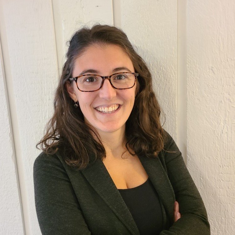 A woman with dark brown, shoulder-length curly hair, wearing glasses, a black top, and a dark blazer, smiling with arms crossed, standing against a white paneled wall.