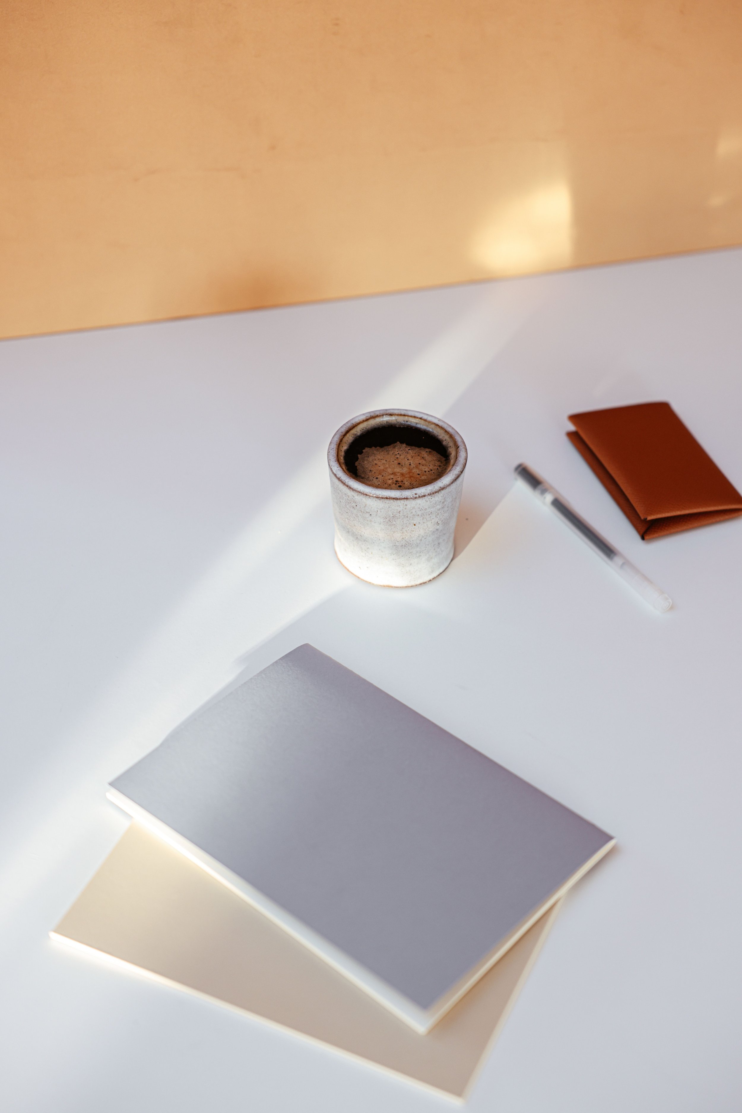A cup of espresso, a pen, a brown notebook, and three closed notebooks in beige, gray, and white, arranged on a white surface with bright lighting.