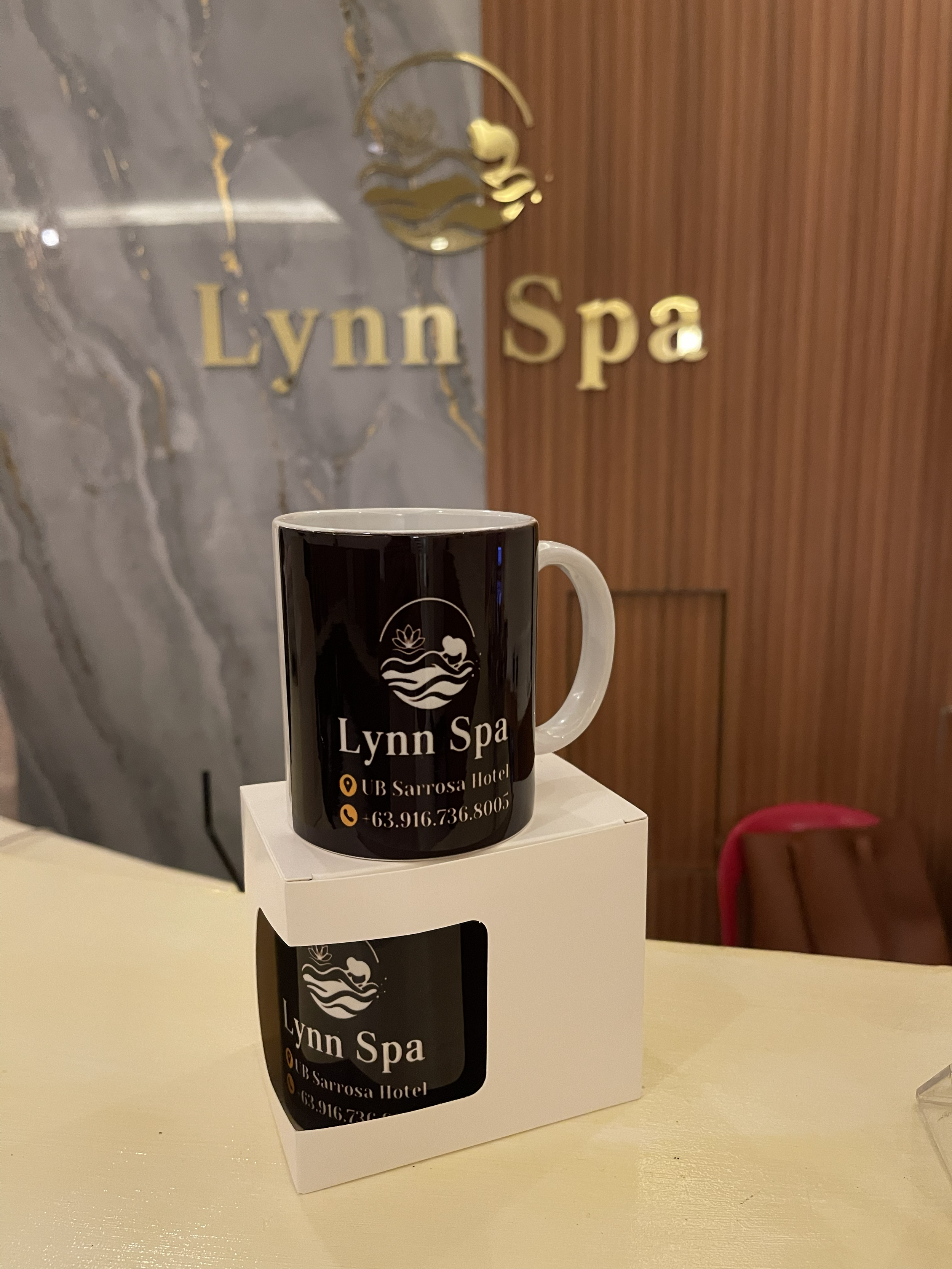 Black mug with white handle on a white stand, displaying the logo and contact information for Lynn Spa at UB Sarrosa Hotel, with a marble wall and wooden panel background.