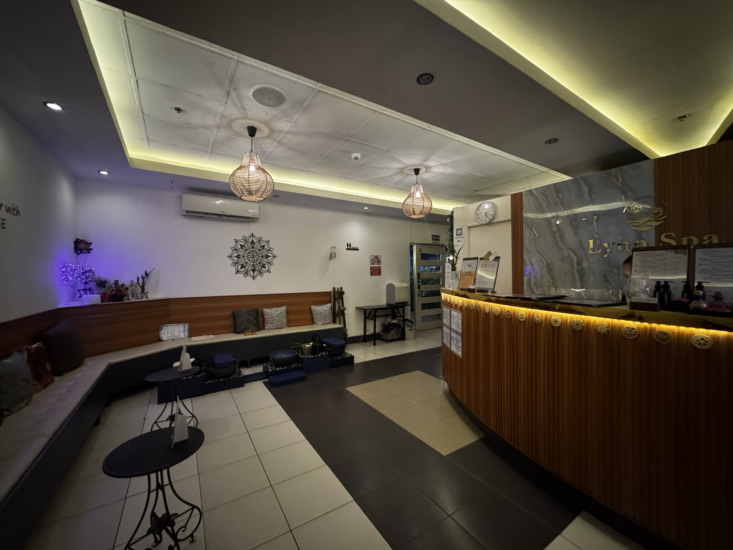 Interior of a restaurant or cafe with a stone wall, a floral wall art, and a reception counter with menu boards. There are hanging pendant lights, seating including a curved sofa with cushions, and small tables with chairs. The ceiling has recessed and ambient lighting.
