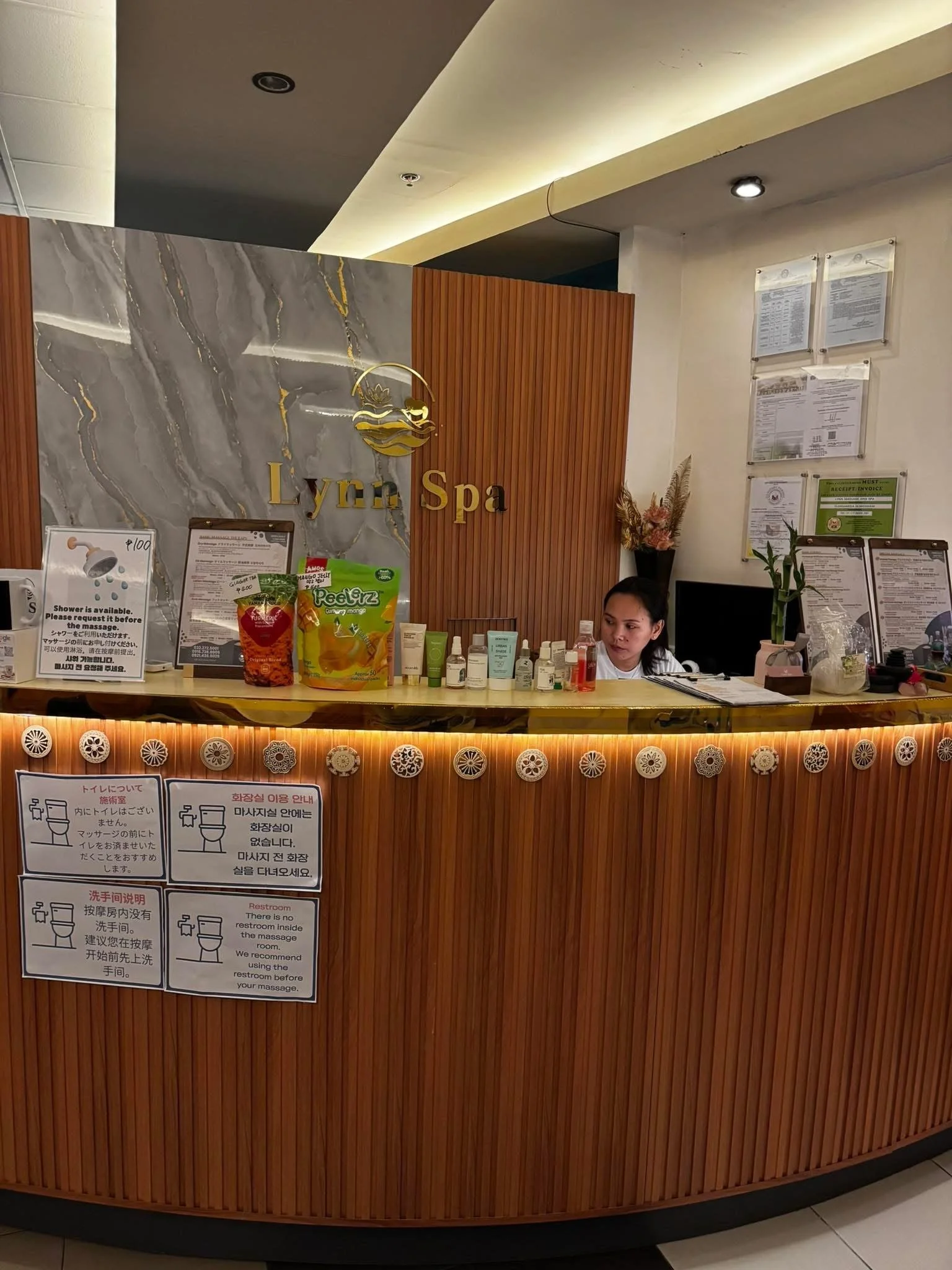 A warm and welcoming reception desk at Lynn Spa Cebu, featuring the brand sign on the wall, a smiling staff member behind the wooden counter, and neatly arranged spa products and brochures.