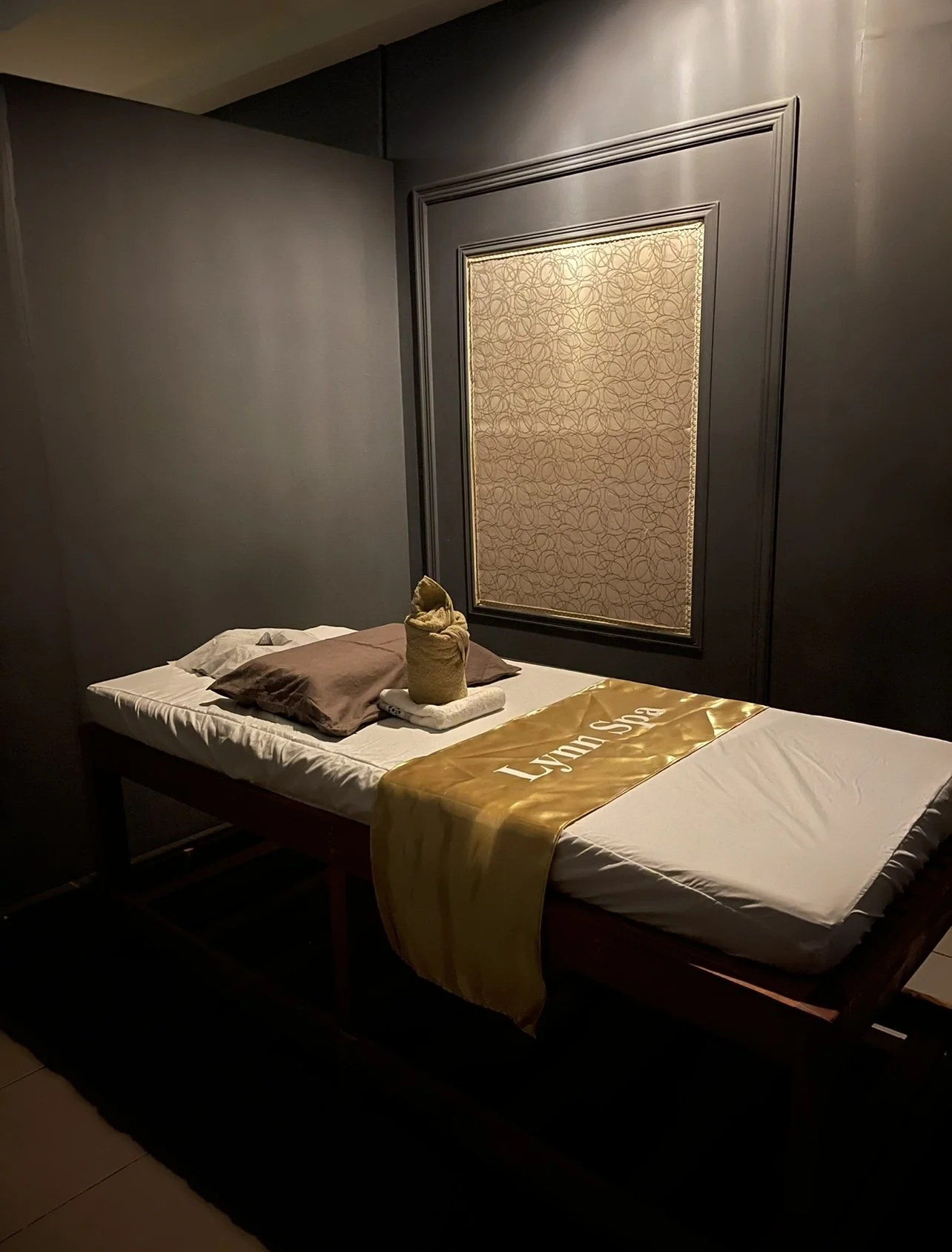 A single bed with white sheets, two brown pillows, and a towel folded into a swan shape on top. There is a folded towel underneath the swan. A yellow bed runner with 'Lynn Sun' written on it is draped across the foot of the bed. The headboard wall features a large decorative pane with a gold pattern, framed with dark molding. The room has dark walls and subdued lighting.