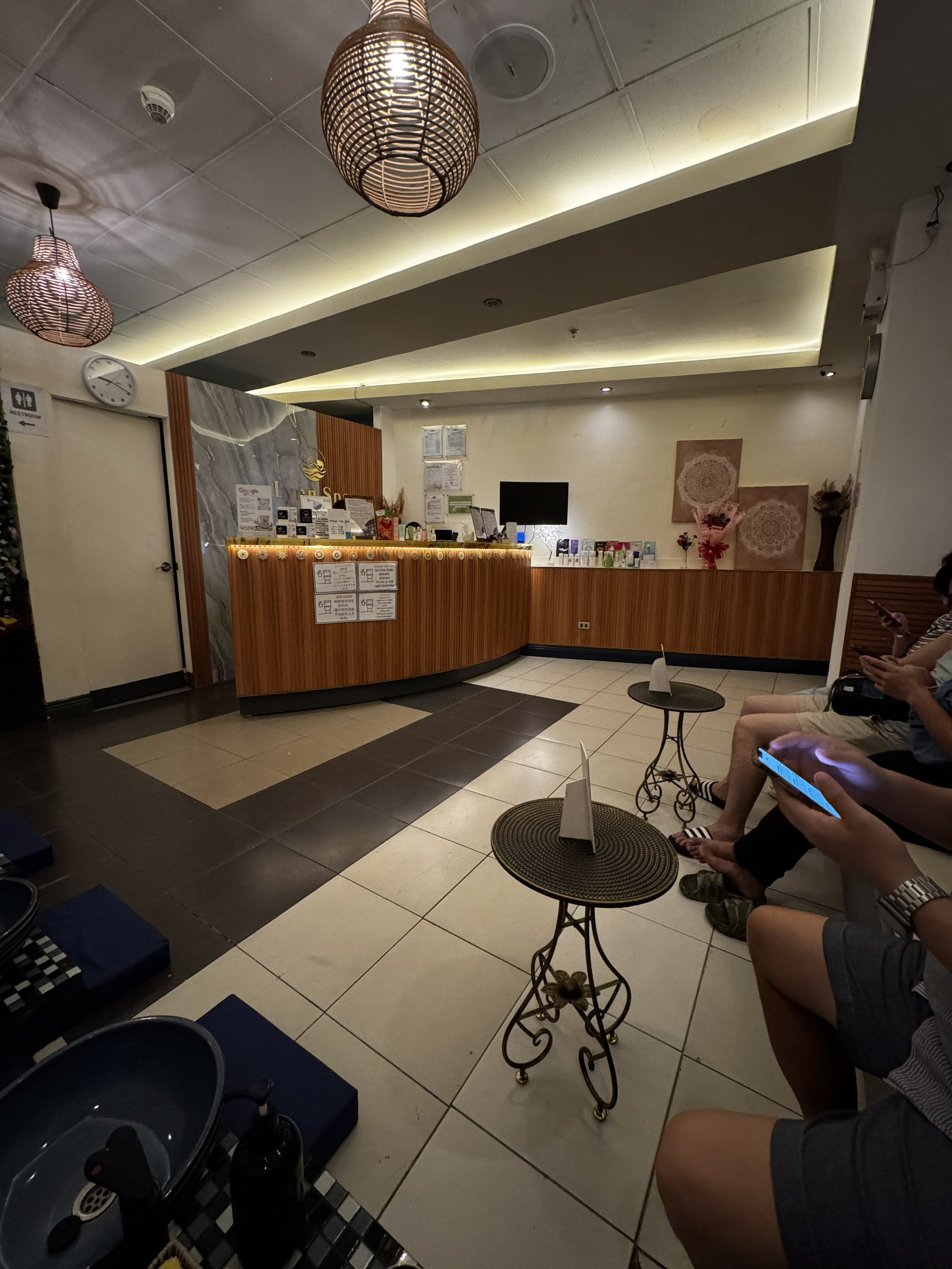 The image shows a reception area with a wooden counter, a small black monitor, various pamphlets or products, and framed artwork on the wall. There are people sitting on chairs, some using their phones. The ceiling has modern lighting fixtures, and there is a clock on the wall indicating the time. The floor is tiled with black and white tiles, and there are small tables and items like water bottles and shoes.