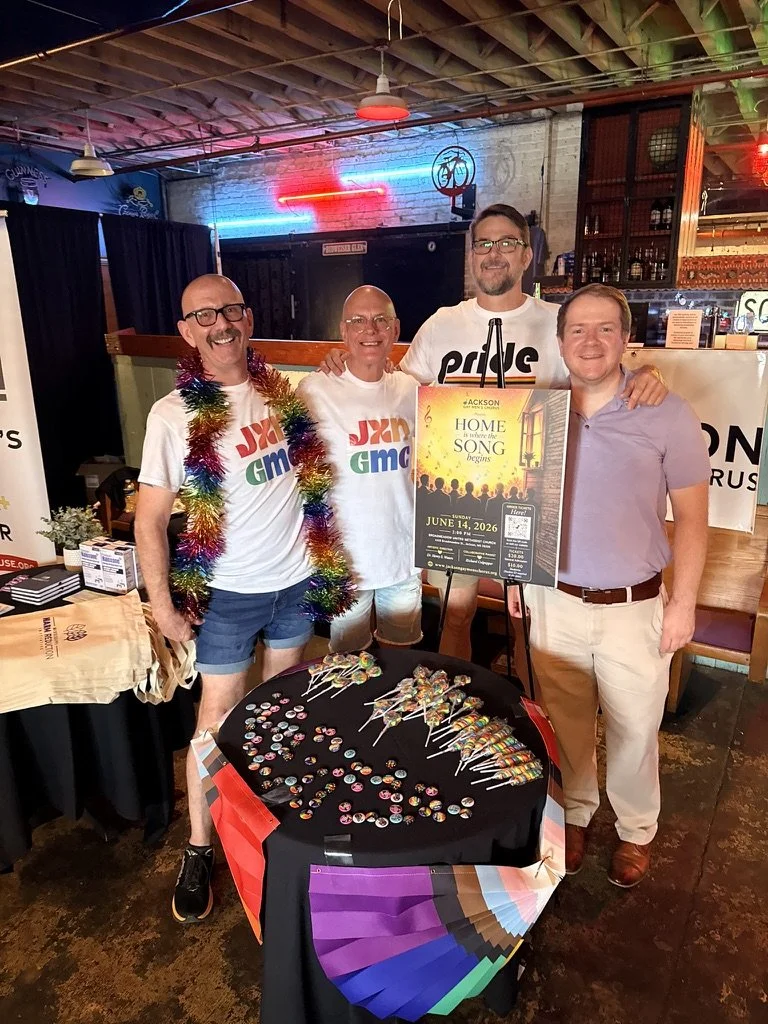 Group of four men standing behind a table with colorful lollipops, pins, and rainbow-themed decorations in a bar or restaurant setting, celebrating an event for 'Jackson Home is there the Song Begins' scheduled for June 14, 2026.