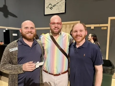 Three men smiling and standing together indoors, with a woman in the background. The man on the left is holding a card, the man in the middle has a rainbow-colored shirt, and the man on the right is wearing a navy polo shirt.