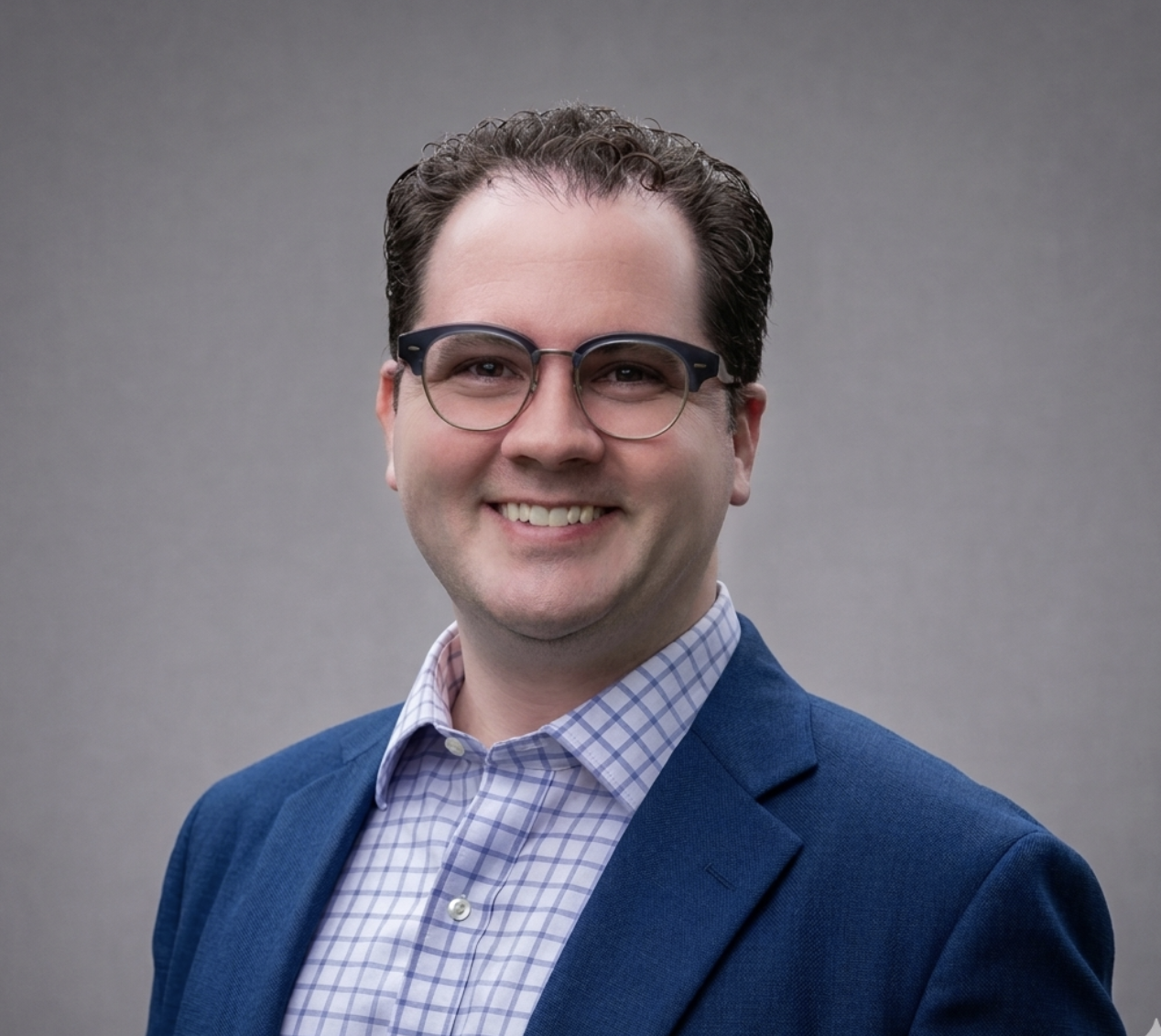 Portrait of a man in glasses smiling, wearing a blue blazer and checkered shirt, against a plain gray background.