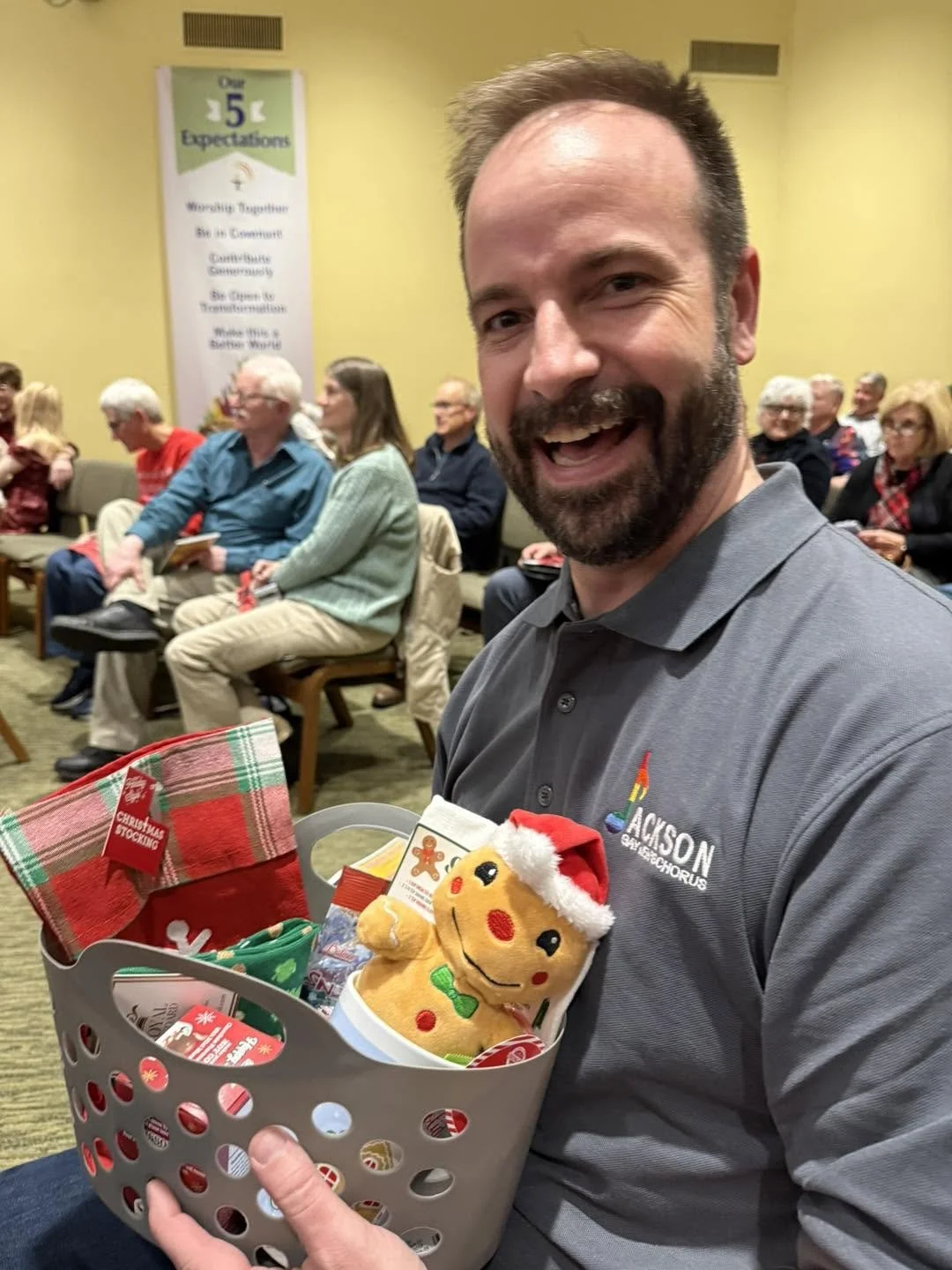 A man smiling and holding a Christmas basket filled with holiday items, wearing a gray polo shirt with a logo, in a room with people seated in the background.