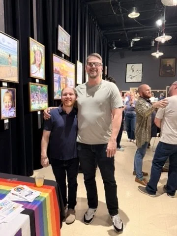 Two men standing in an art gallery, smiling, with paintings on the wall behind them. One is shorter with a beard, wearing a dark shirt, and the taller is wearing glasses and a light-colored T-shirt. There are other people in the background and a rainbow flag on a table.