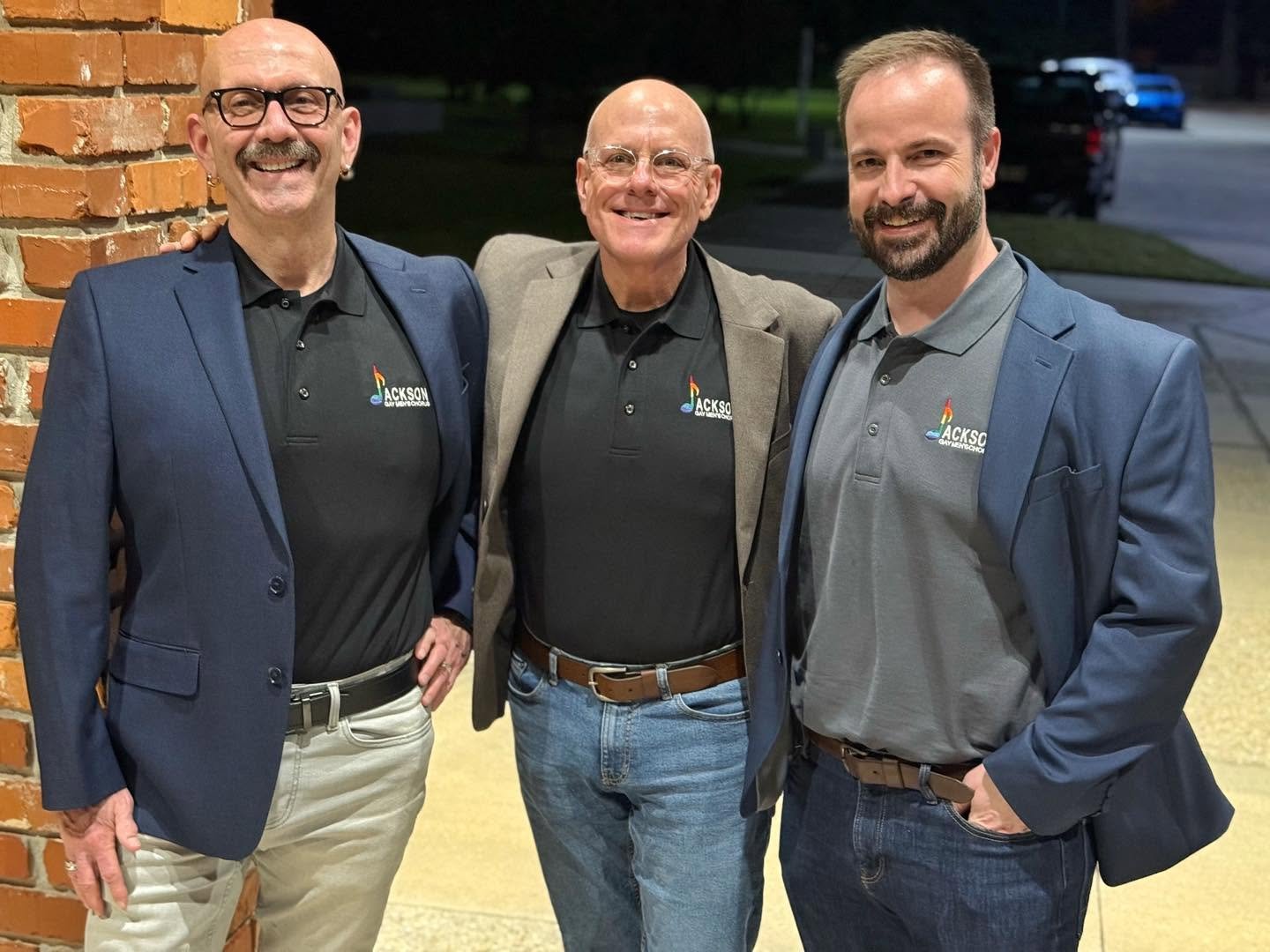 Three men standing together in front of a brick wall and outdoors at night, wearing blazers and polo shirts with `Jackson Gay Men’s Chorus` logo, smiling for the camera.