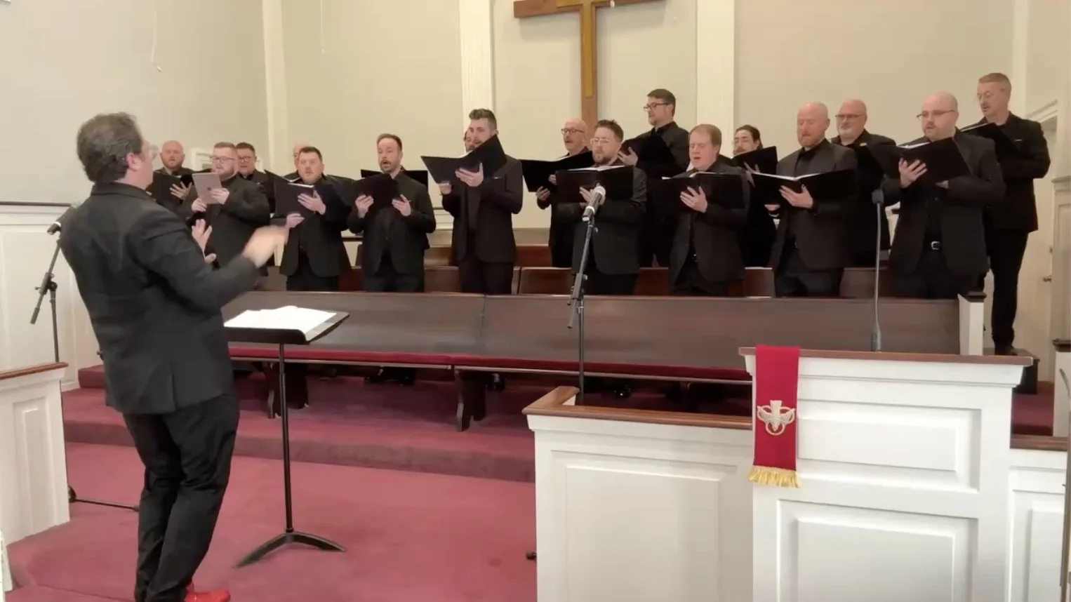 A male choir dressed in black suits singing on a church stage, guided by a conductor standing in front of them, with a cross hanging on the wall behind.