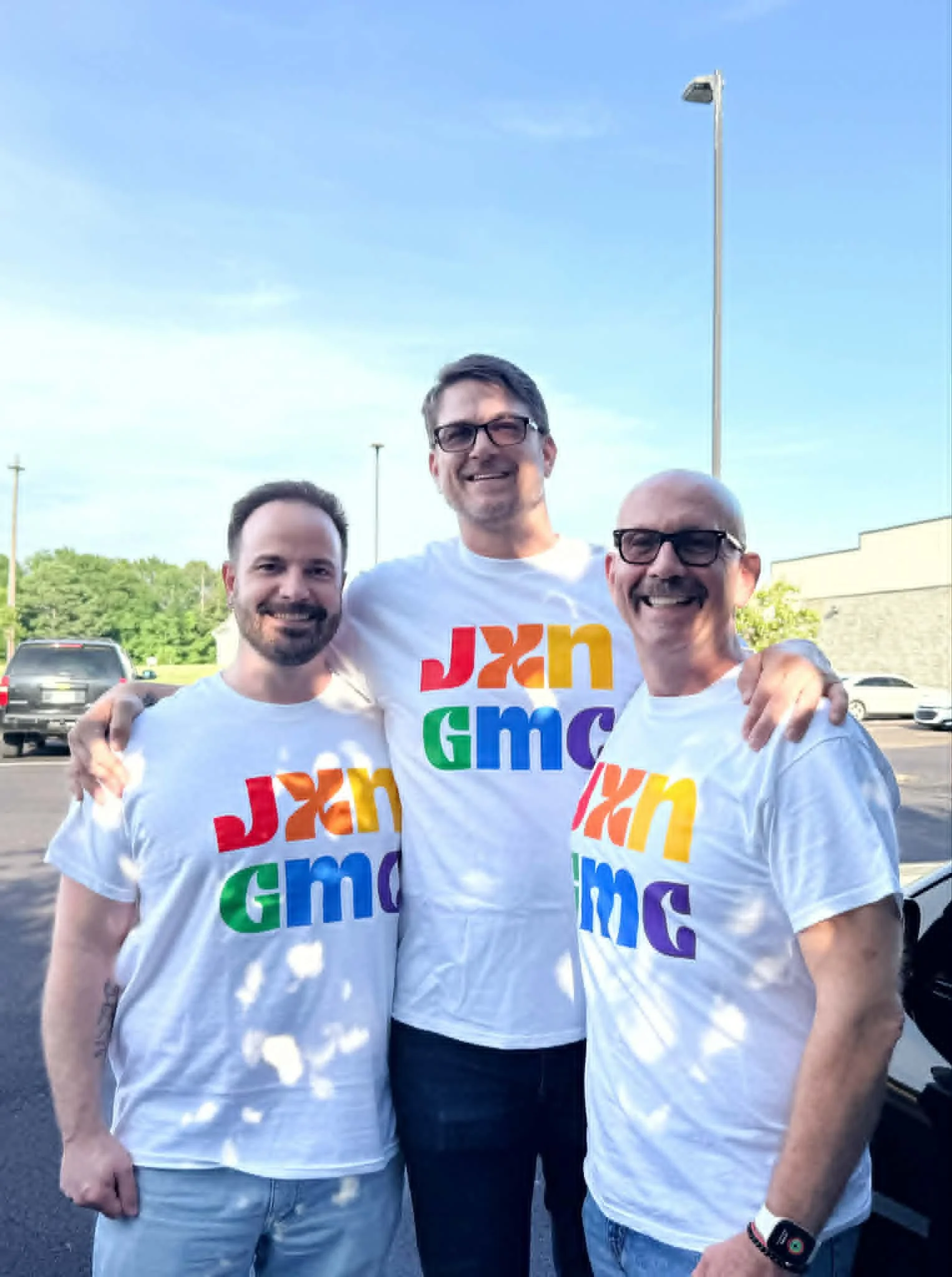 Three men standing outside in a parking lot, smiling and wearing white T-shirts with colorful text. The background includes trees, parked cars, a building, and a tall streetlamp. The sky is partly cloudy.