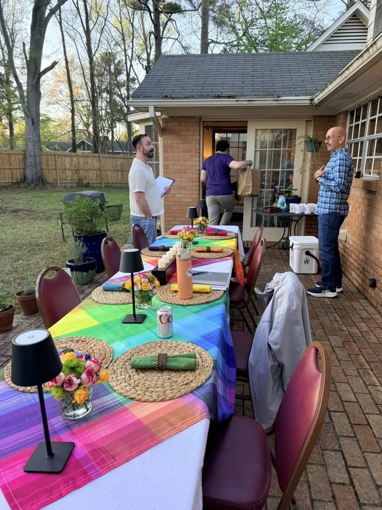 Backyard patio with a long dining table covered in a rainbow-colored tablecloth, decorated with small black lamps, patterned placemats, flowers, rolled napkins, and drinks, set for a gathering. Three men are present, one opening the door into the house, another holding a notepad, and the third standing with arms crossed, near brick house with a fenced yard and trees in the background.