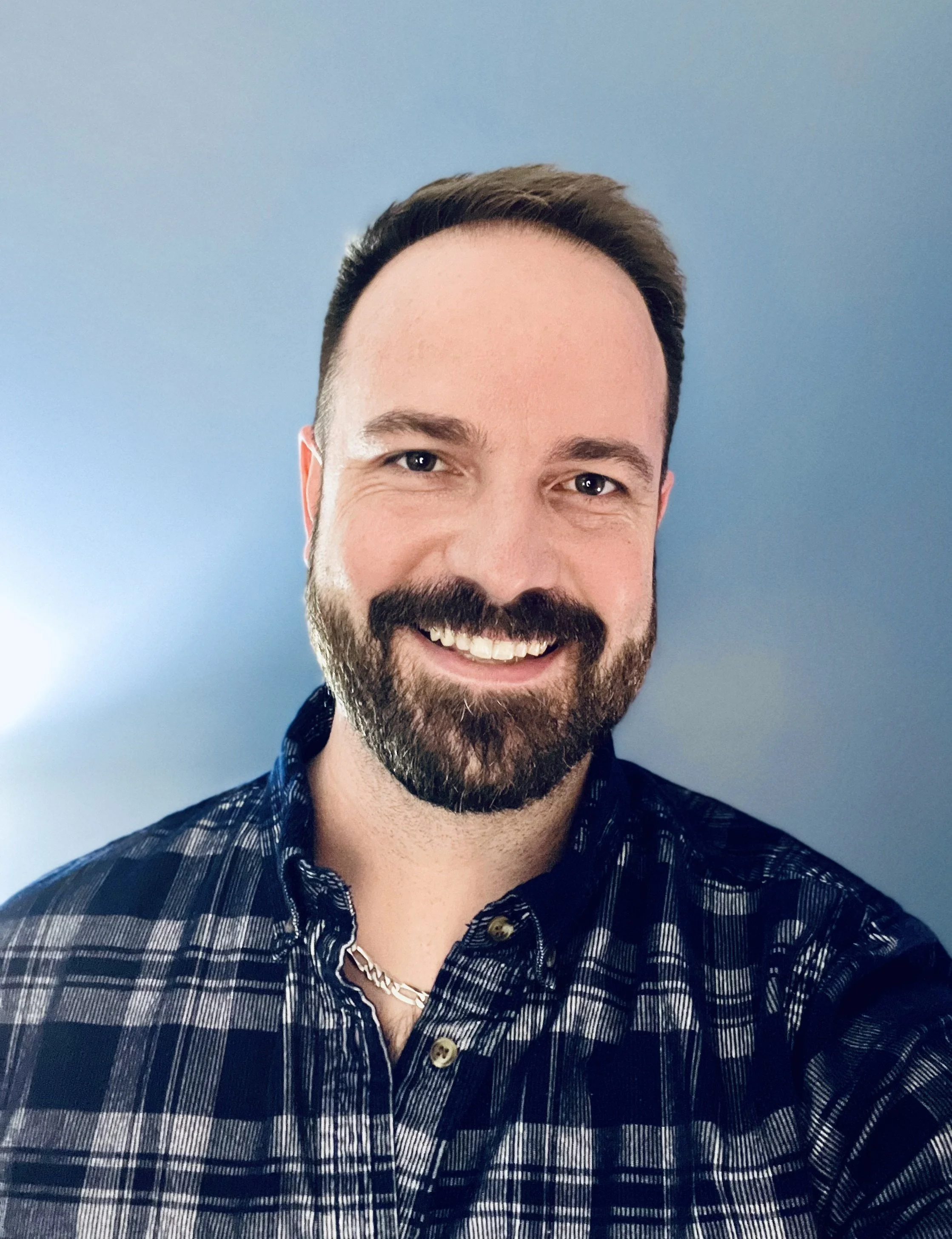 Close-up of a smiling man with brown hair and a beard, wearing a black and white plaid shirt, against a plain blue background.