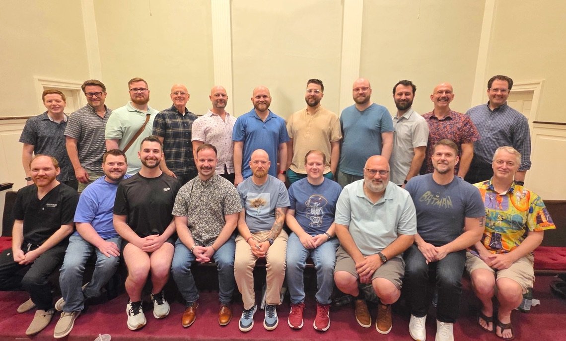 Group of 18 men posing for a photo in a large room with light green walls.