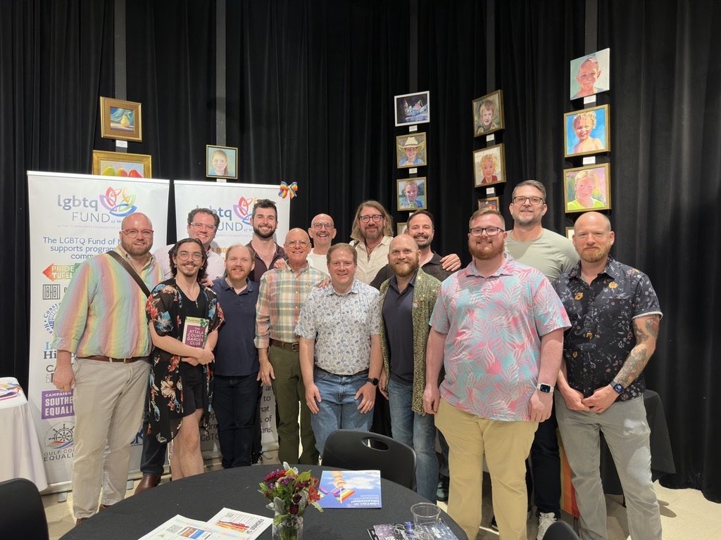 Group of people posing together at an event with LGBTQ+ support banners, art pieces on black curtains in the background, and a table with flowers and brochures in the foreground.