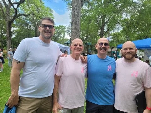 Four people standing together outdoors in a park, smiling, with tents and trees in the background. They are wearing casual t-shirts in light colors.
