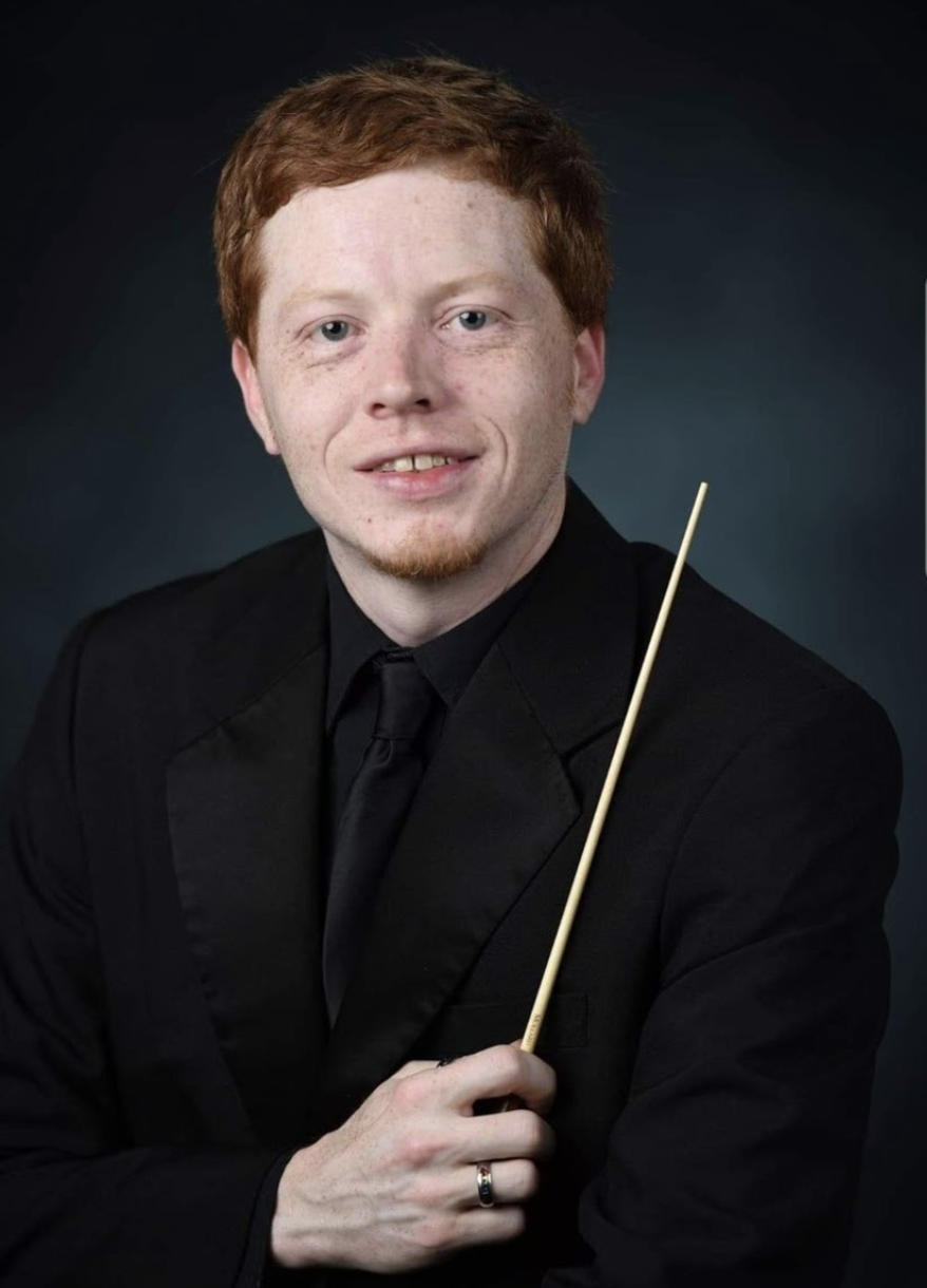 Young man with red hair, freckles, and a goatee in a black suit and tie, holding a conductor's baton.
