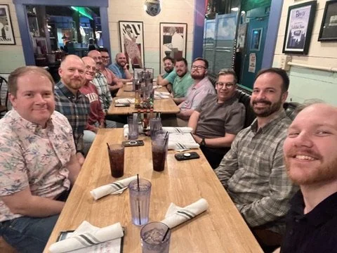 A group of fifteen men sitting around a long wooden table in a restaurant, smiling at the camera.