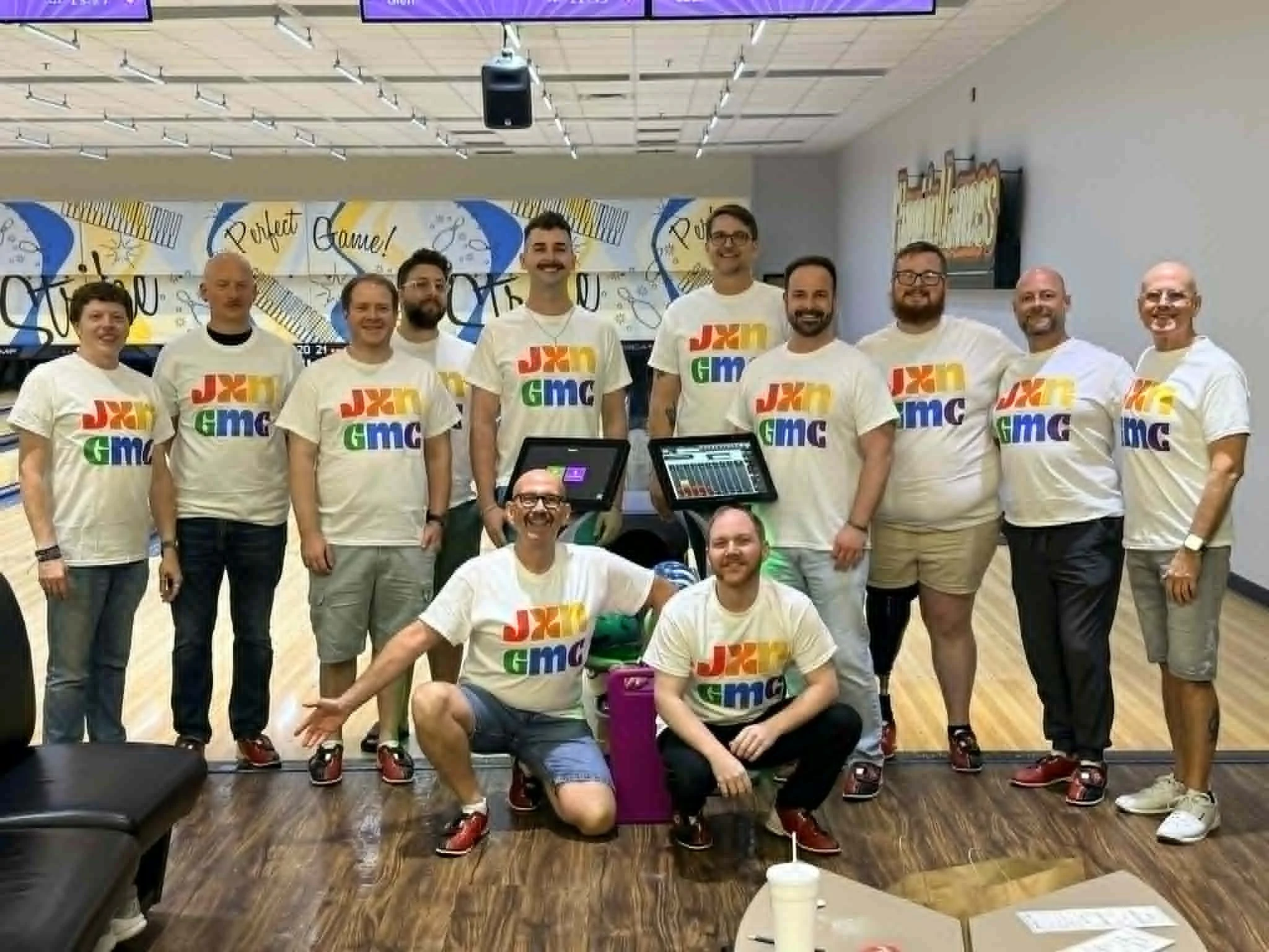 A group of people standing together in a room, all wearing white T-shirts with colorful logos, smiling and looking at the camera during an event or gathering.