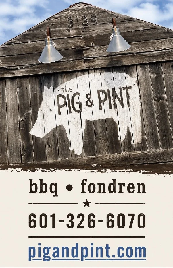 Sign for The Pig & Pint restaurant with a pig silhouette, featuring BBQ and fondu, phone number 601-326-6070, and website pigandpint.com, mounted on a wooden barn with metal barn lights above, under a partly cloudy sky.