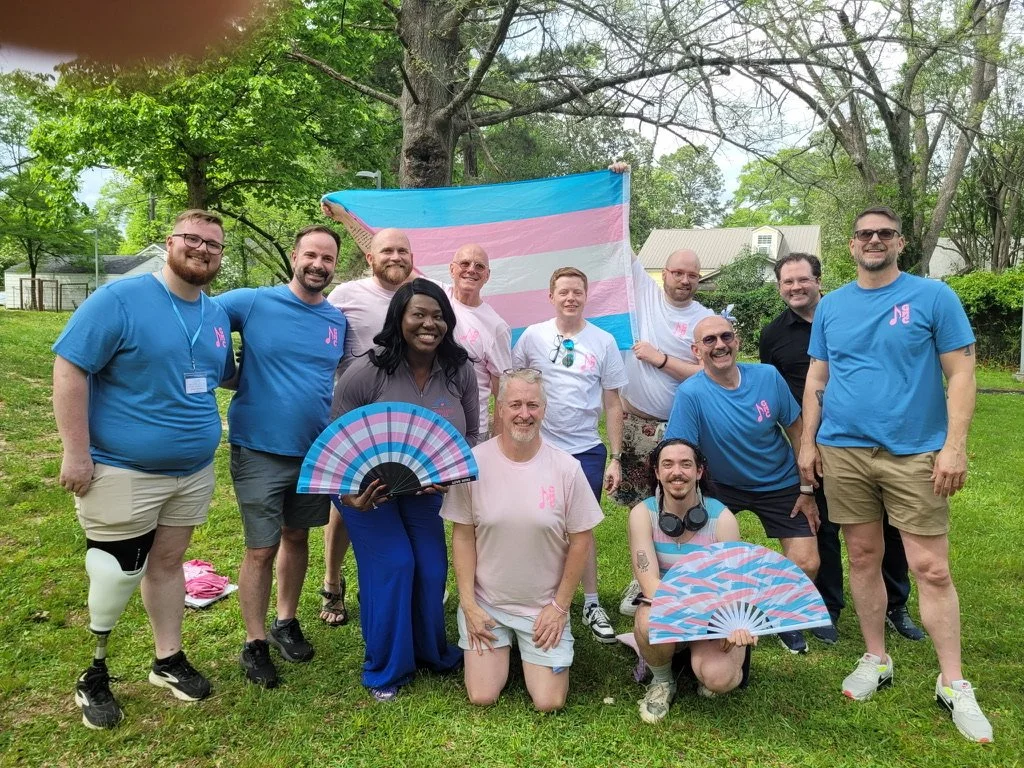 Group of people celebrating LGBTQ+ pride outdoors, holding a transgender pride flag and pink, blue, and white fans.