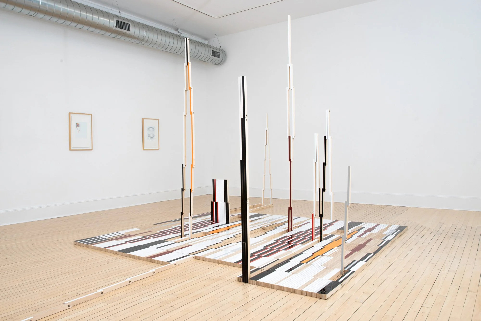An art installation with vertical black, white, and red striped poles of varying heights on a multicolored striped base, set in a gallery with white walls and light hardwood floors.