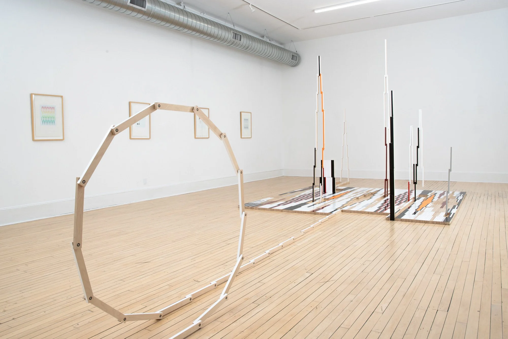 An art installation in a gallery with wooden flooring, consisting of vertical rods in black, white, orange, and red, arranged in a pattern resembling a city skyline, with framed geometric artwork on white walls and a white wooden frame structure in the foreground.