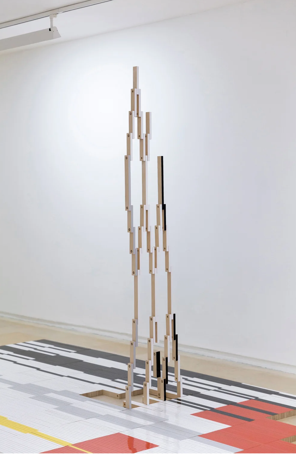 An abstract wooden sculpture made of multiple vertically aligned wooden planks connected with hinges, assembled in a tall, irregular pattern, placed on a geometric-patterned floor with gray, black, red, and yellow accents.