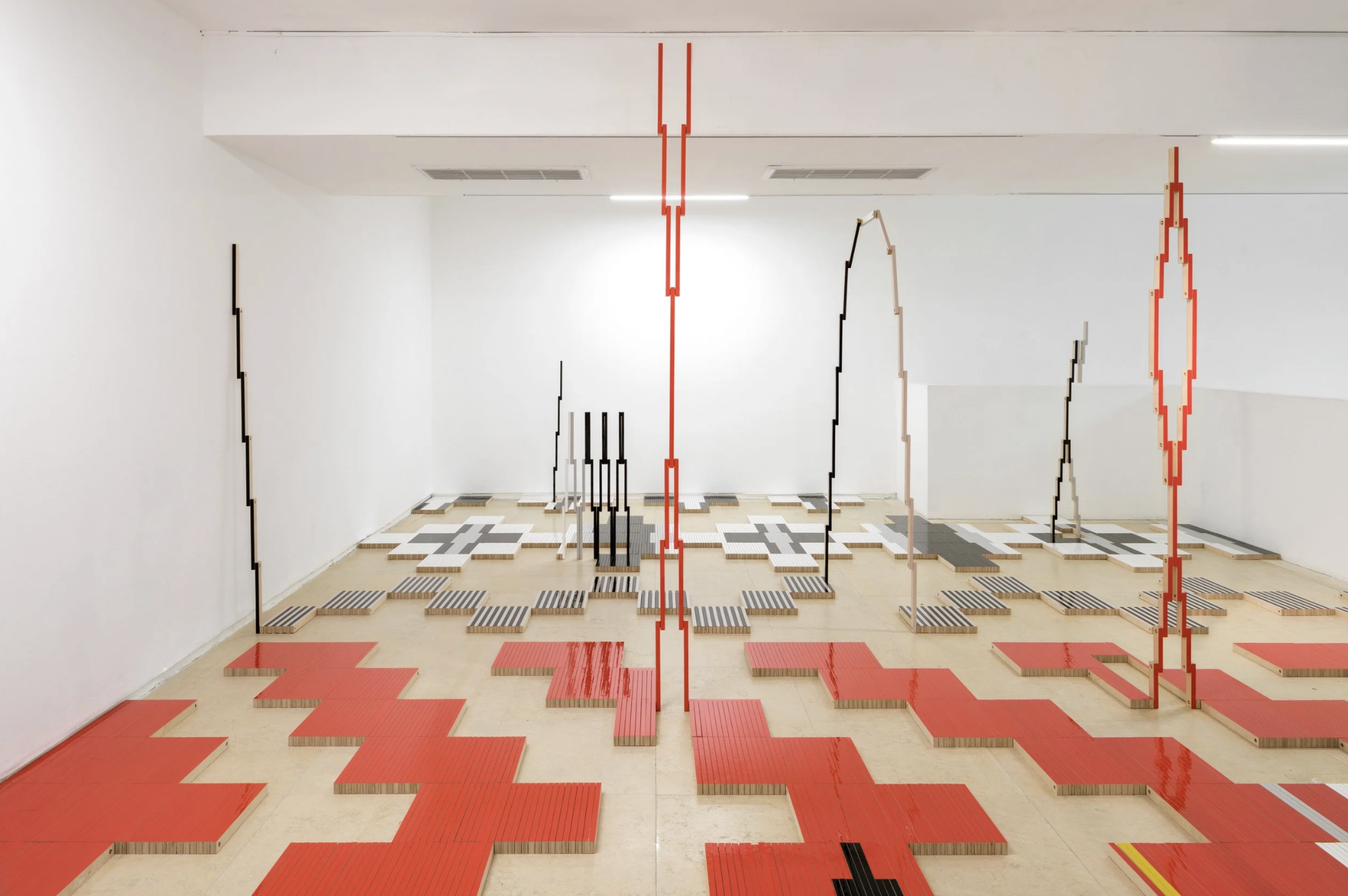 An art installation with various tall, vertical structures made of wooden planks and metal, painted in black, red, and beige, arranged in a pattern on a tiled floor in a white gallery space.