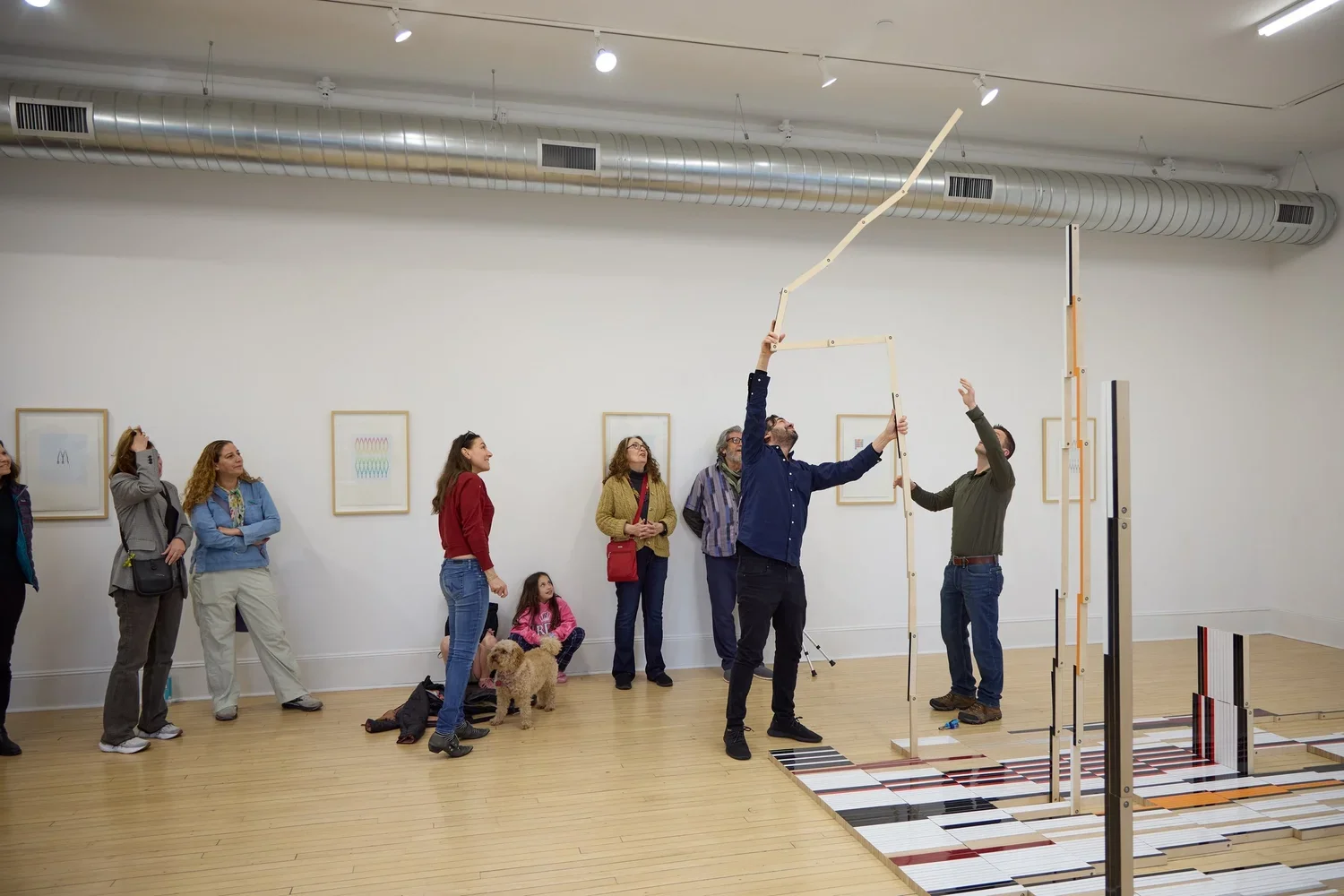 People gathered in an art gallery, observing a kinetic sculpture being assembled or explained by two men, with colorful panels on the floor and framed artwork on the walls.