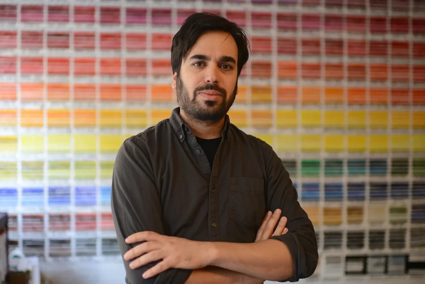 Man with dark hair and beard standing with arms crossed in front of a wall of colorful paint samples.