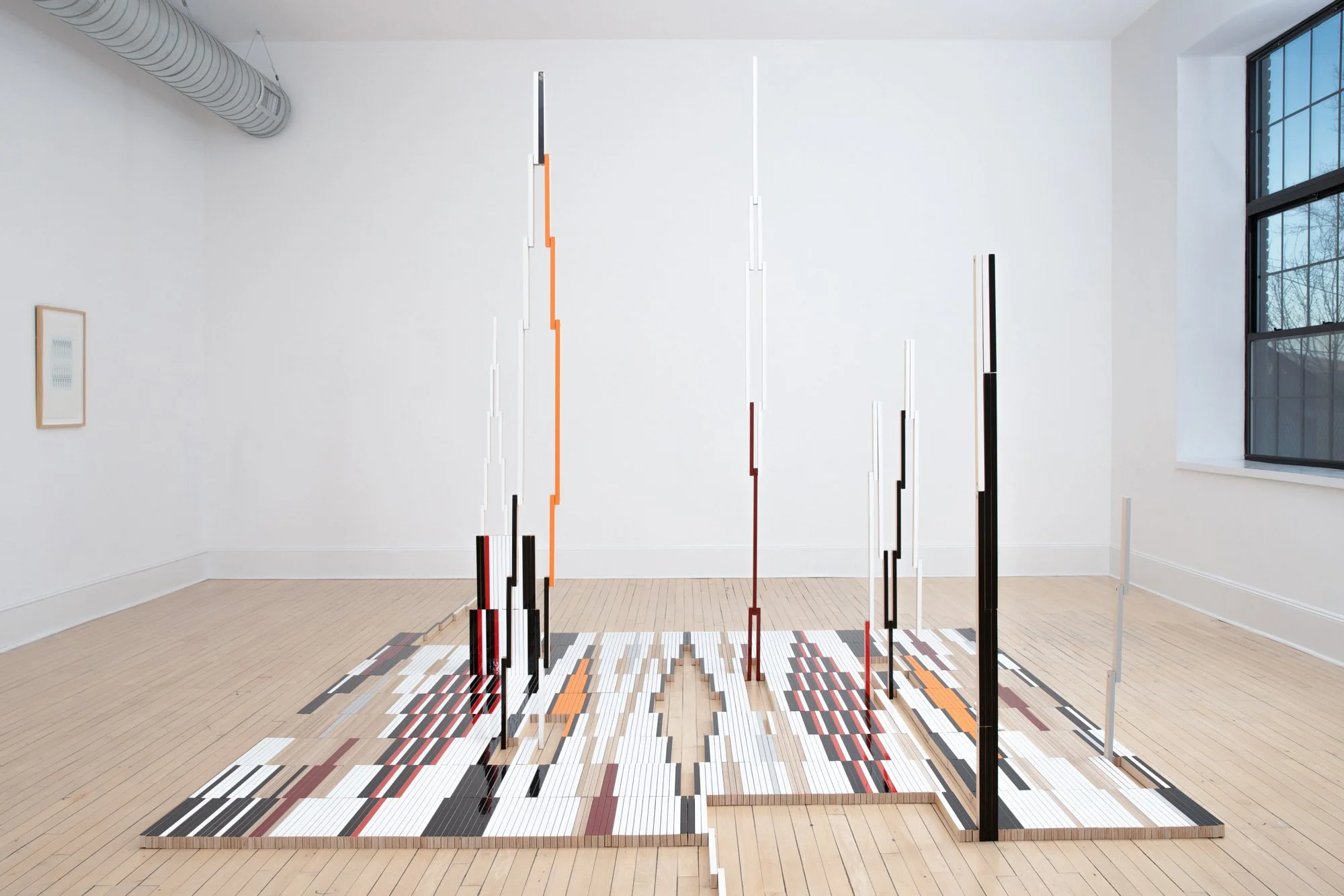 An art installation features vertical striped poles of various heights arranged over a multicolored tile floor in a gallery with white walls and large windows.