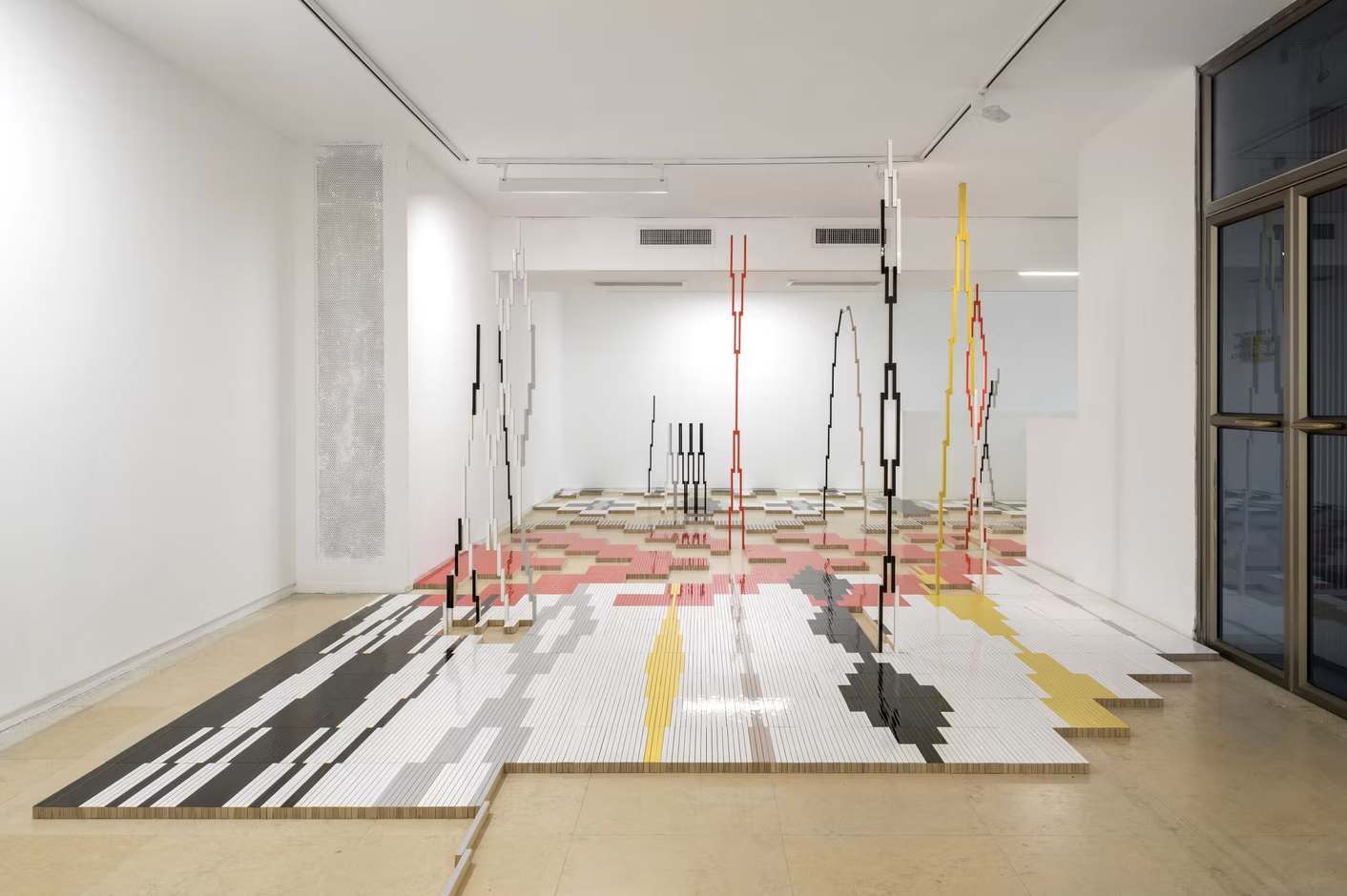 Modern art installation with vertical colored and black structures on a pixelated patterned floor in a white gallery space.