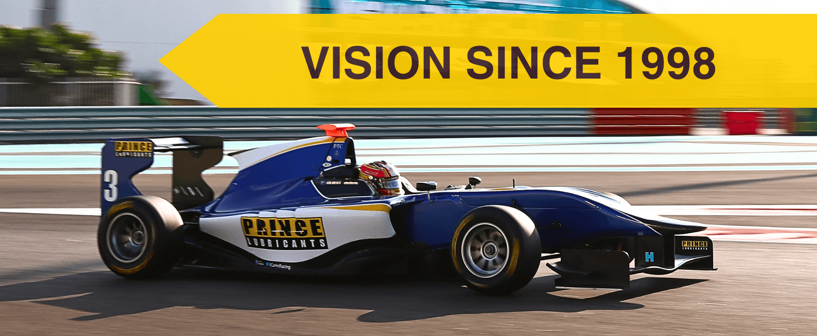 A race car on a track with a yellow sign above it that reads 'Vision Since 1998'. The car has Prince Lubricants branding and is blue, white, and black with the number 3 on the rear wing.
