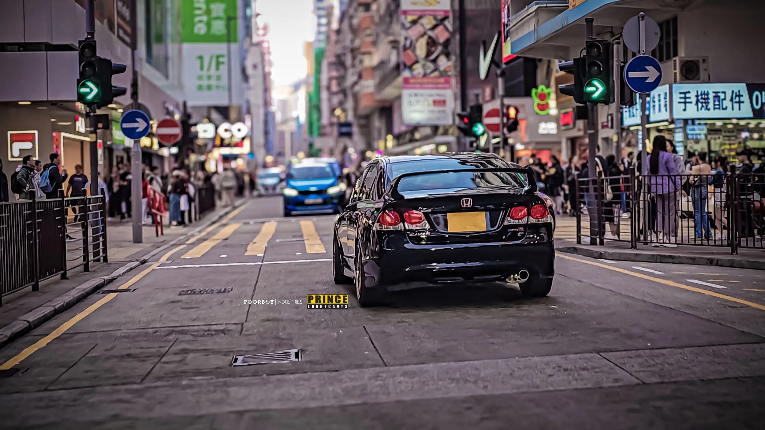 A busy city street with cars, pedestrians, and illuminated signs. A black Honda car is in foreground with green traffic light indicating go. People are waiting and walking along sidewalks. Bright advertisements and signs are visible on buildings.