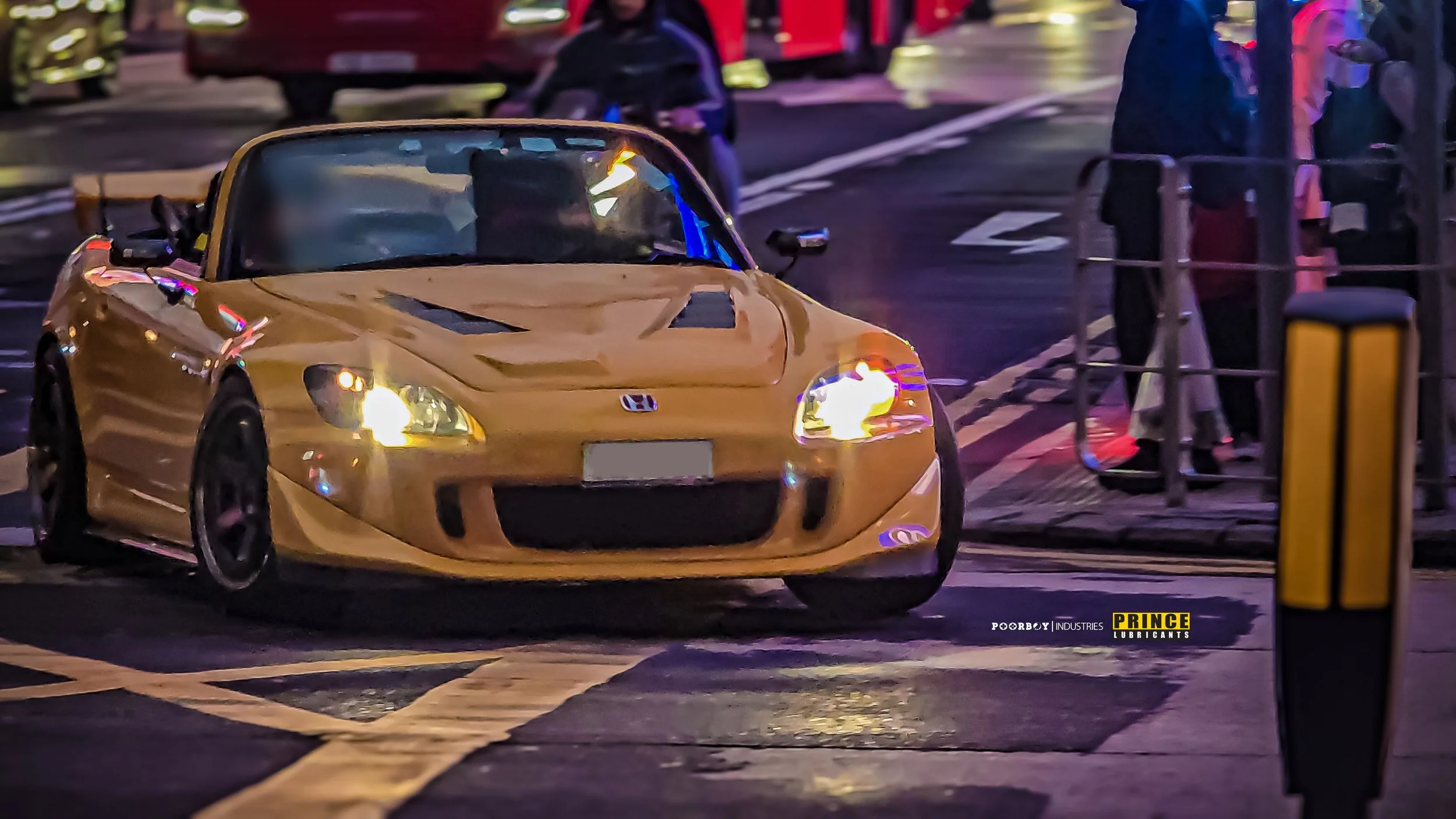 A yellow Honda S2000 sports car with a damaged front end, including a crushed hood, parked at a parking lot entrance at night. Several people are standing nearby, and there are cars and a barrier gate in the background.