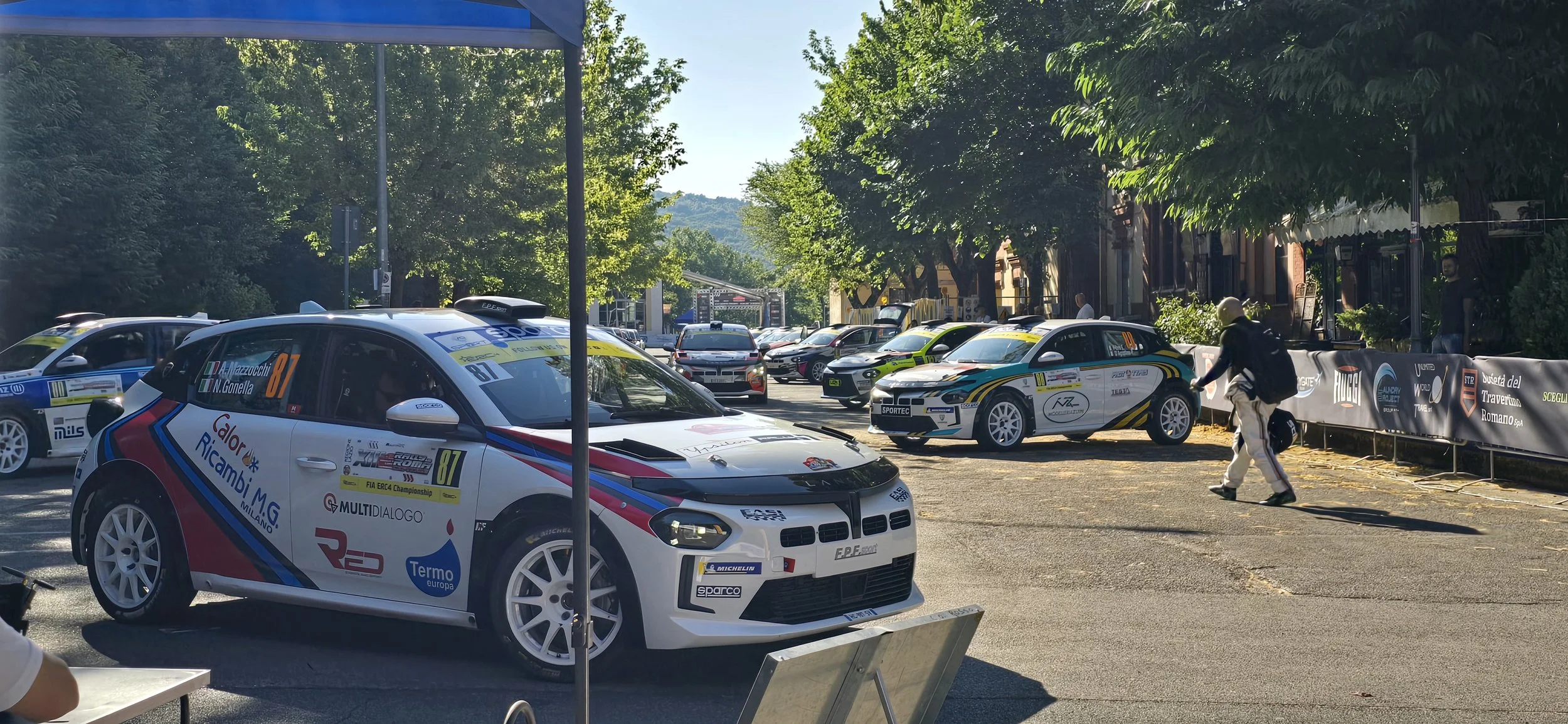 Race cars parked in a lineup on a tree-lined street during daytime, with people walking nearby and spectators in the background.