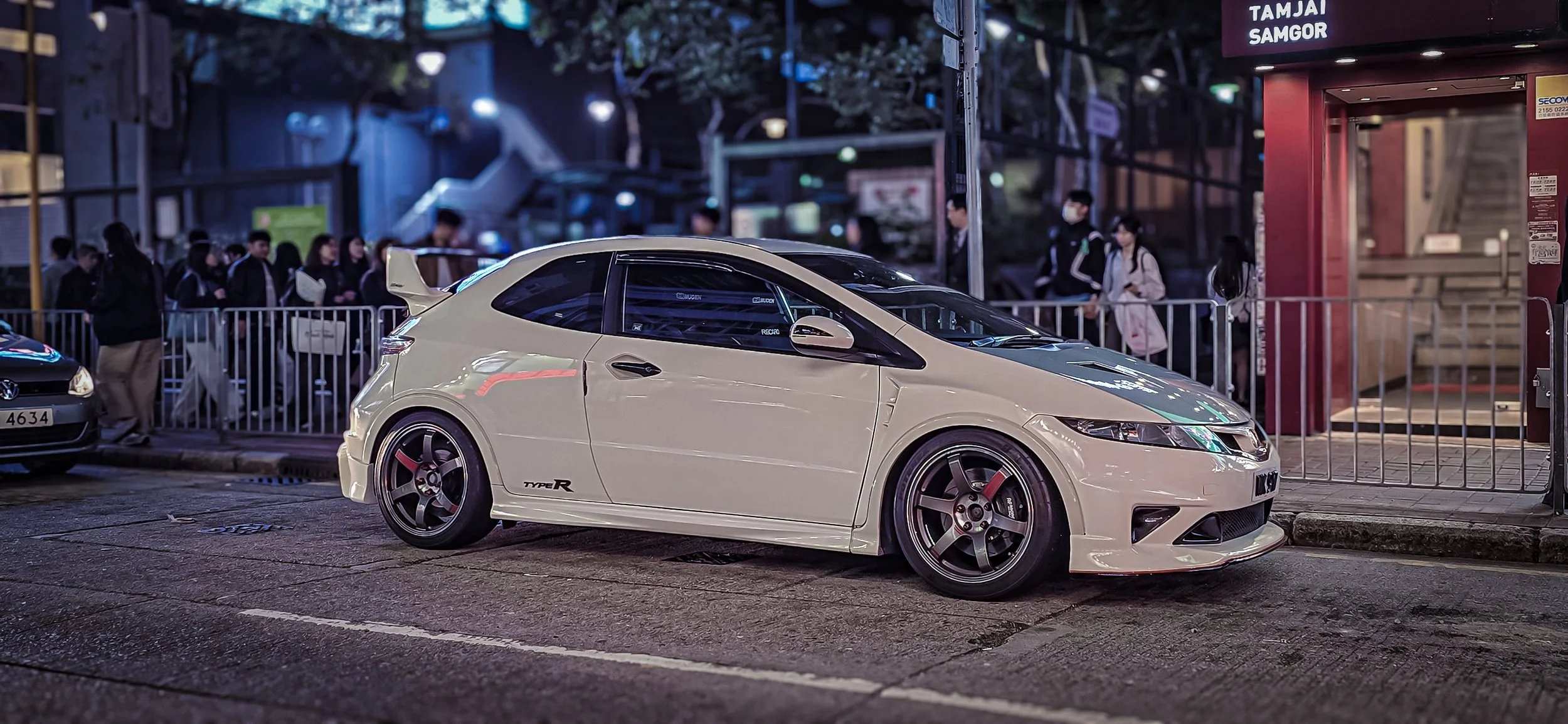 White sporty car with a spoiler parked on the street at night, with people waiting behind a metal fence and a red elevator nearby.