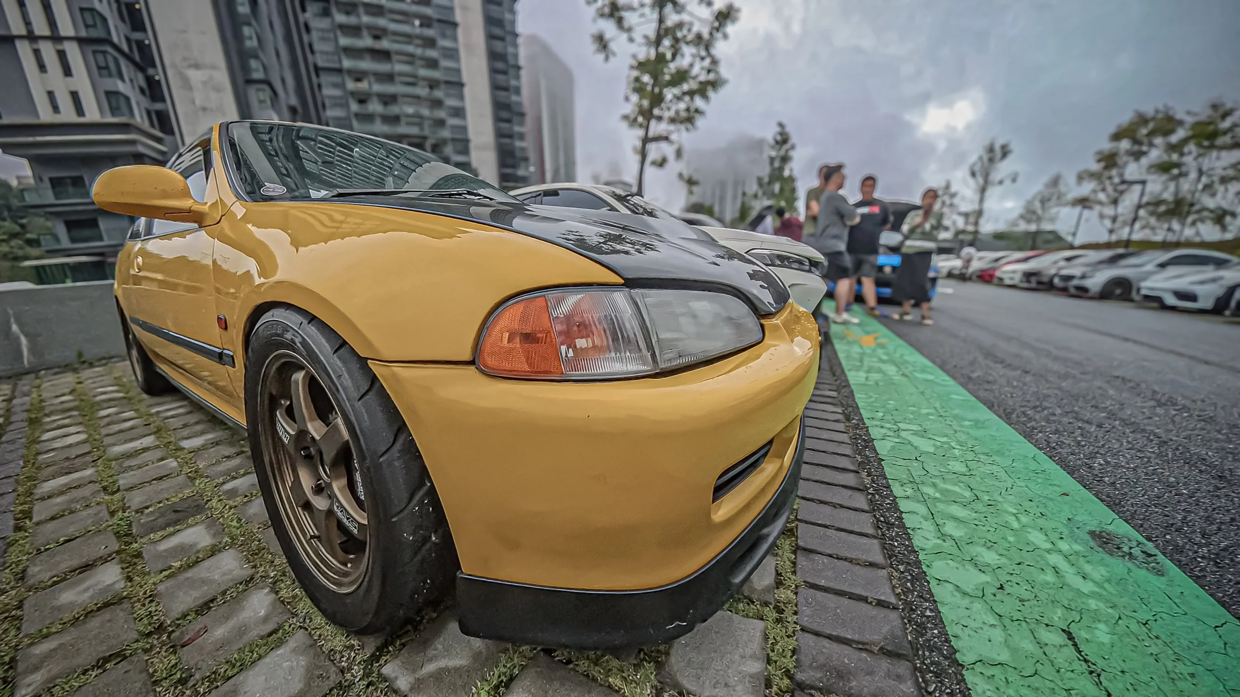 A yellow sports car with a black hood parked in a city parking lot, with people standing near other cars in the background and tall buildings around.