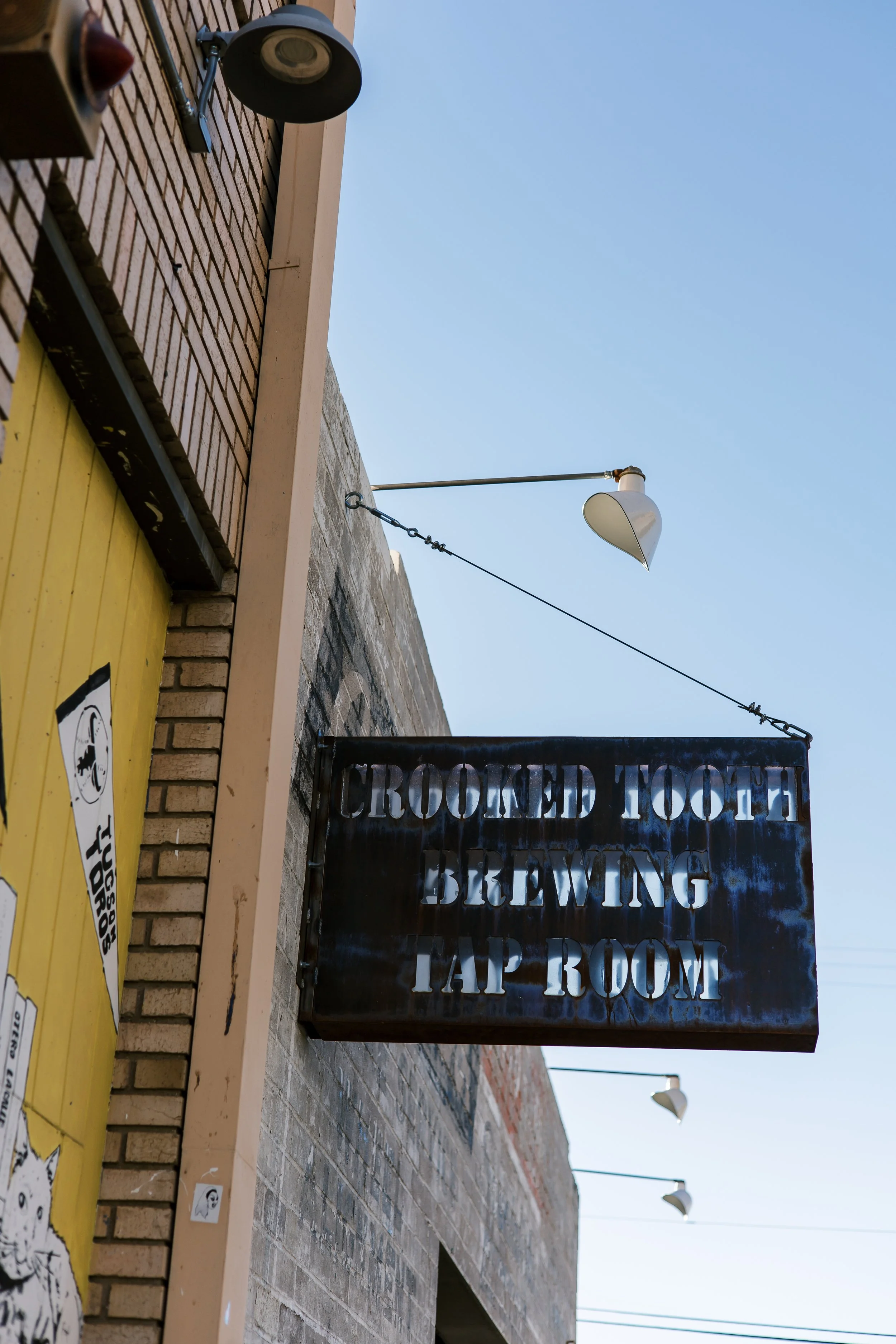 Sign hanging outside a building with the words "Crooked Tooth Brewing Tap Room." The building has a brick exterior with outdoor lighting fixtures and a yellow painted section.
