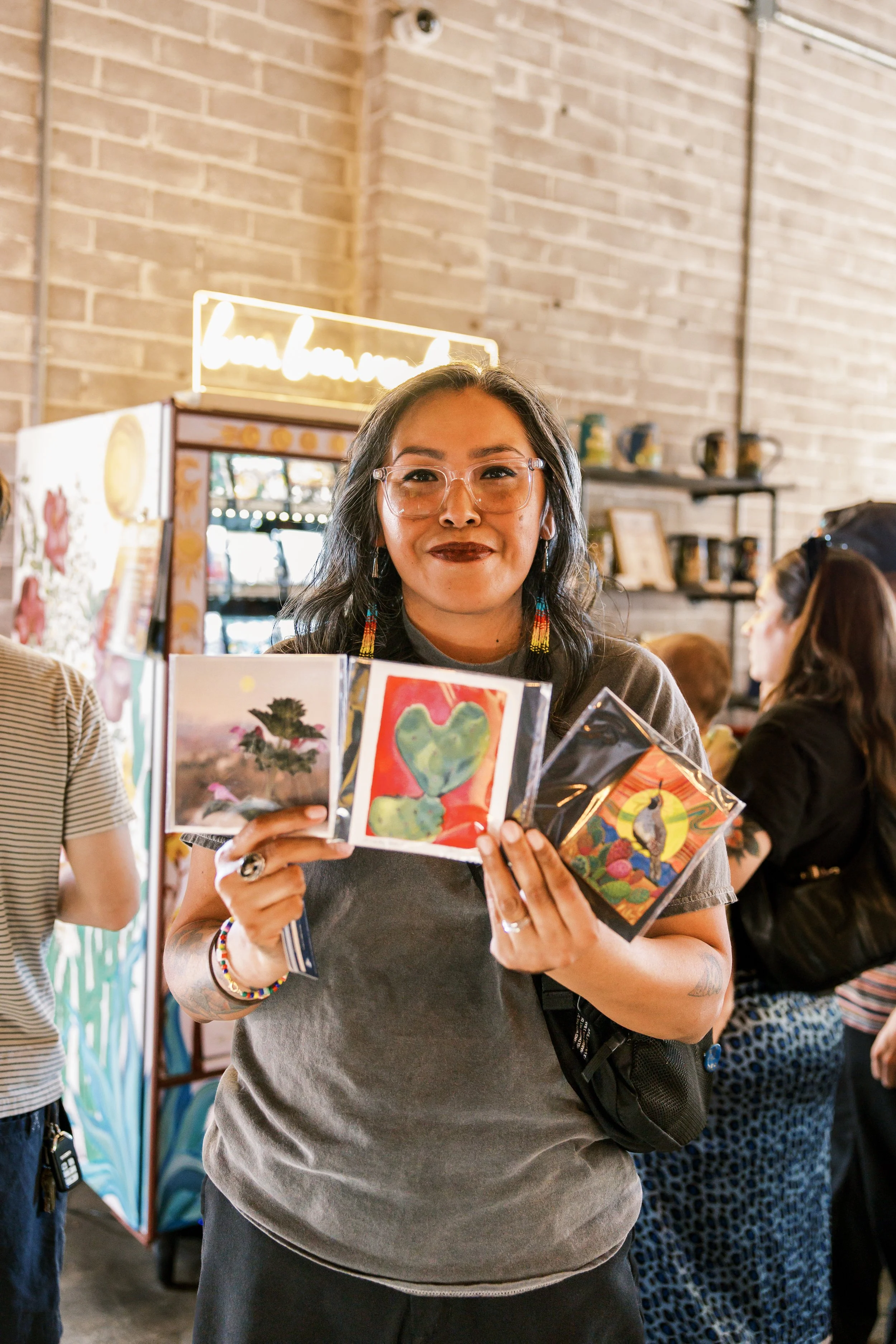 Woman with glasses holding art prints in a gallery or craft fair. Background shows brick wall and other people browsing art.