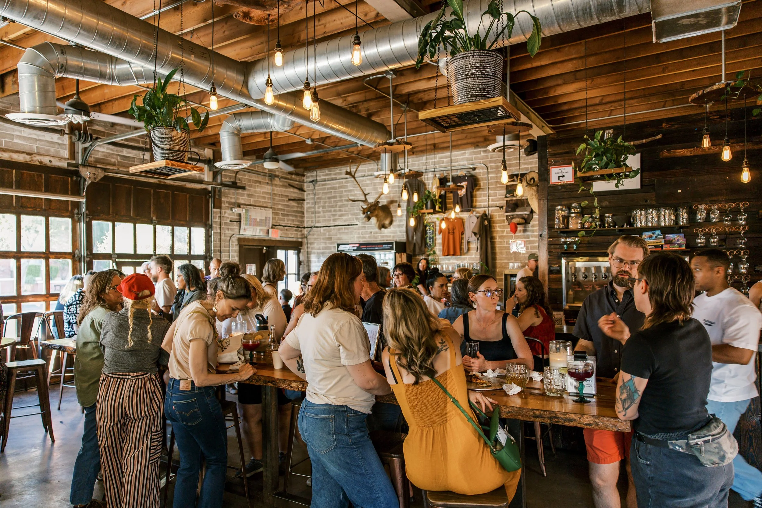 A bustling indoor restaurant or bar with exposed brick and wood decor, filled with a diverse group of people socializing, some standing and some seated, with warm lighting and decorative plants hanging from the ceiling.