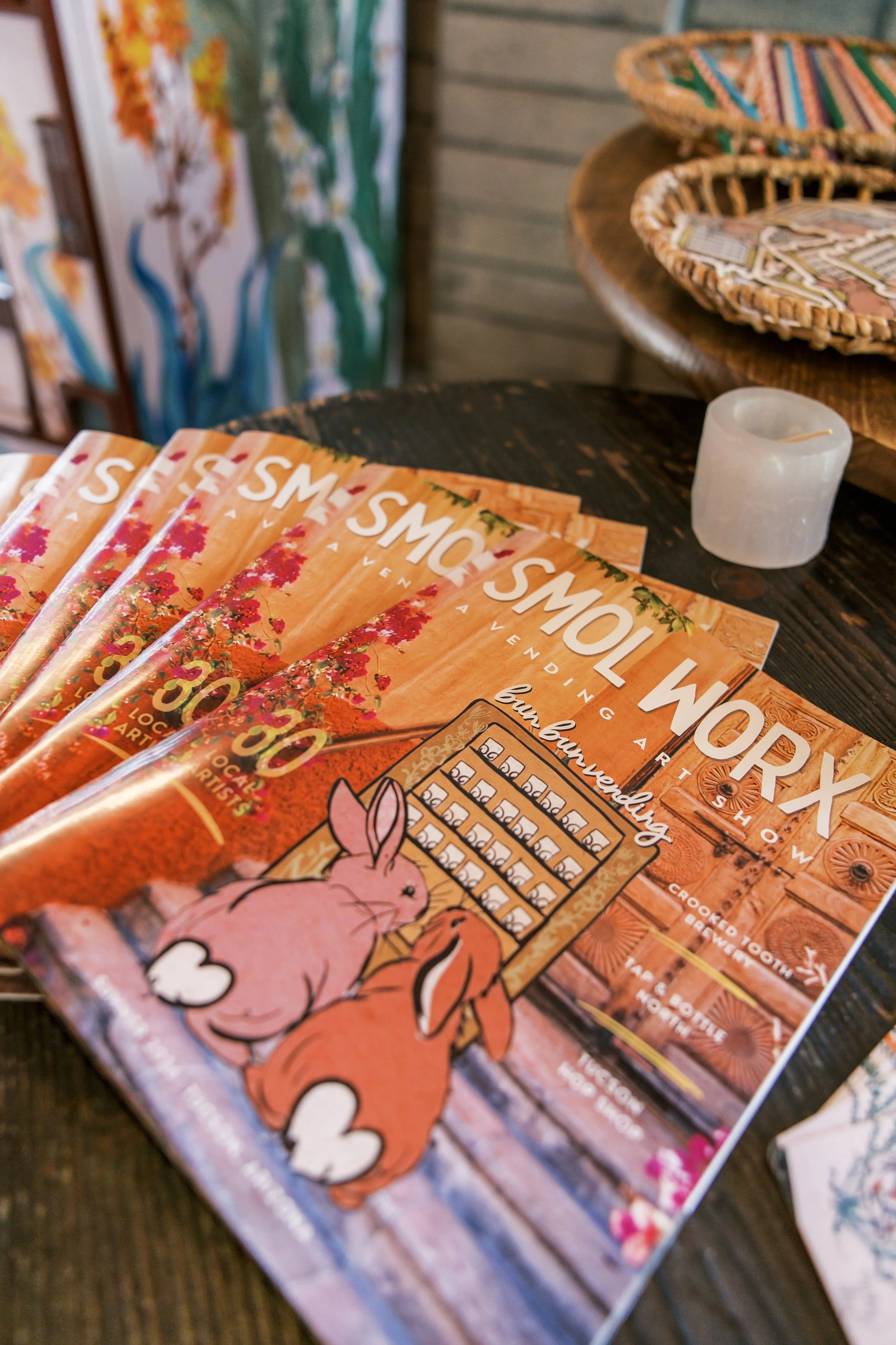 A stack of flyers advertising the Smol Worx event with a pink cartoon bunny rabbit in the foreground, placed on a wooden table with woven baskets and artwork in the background.