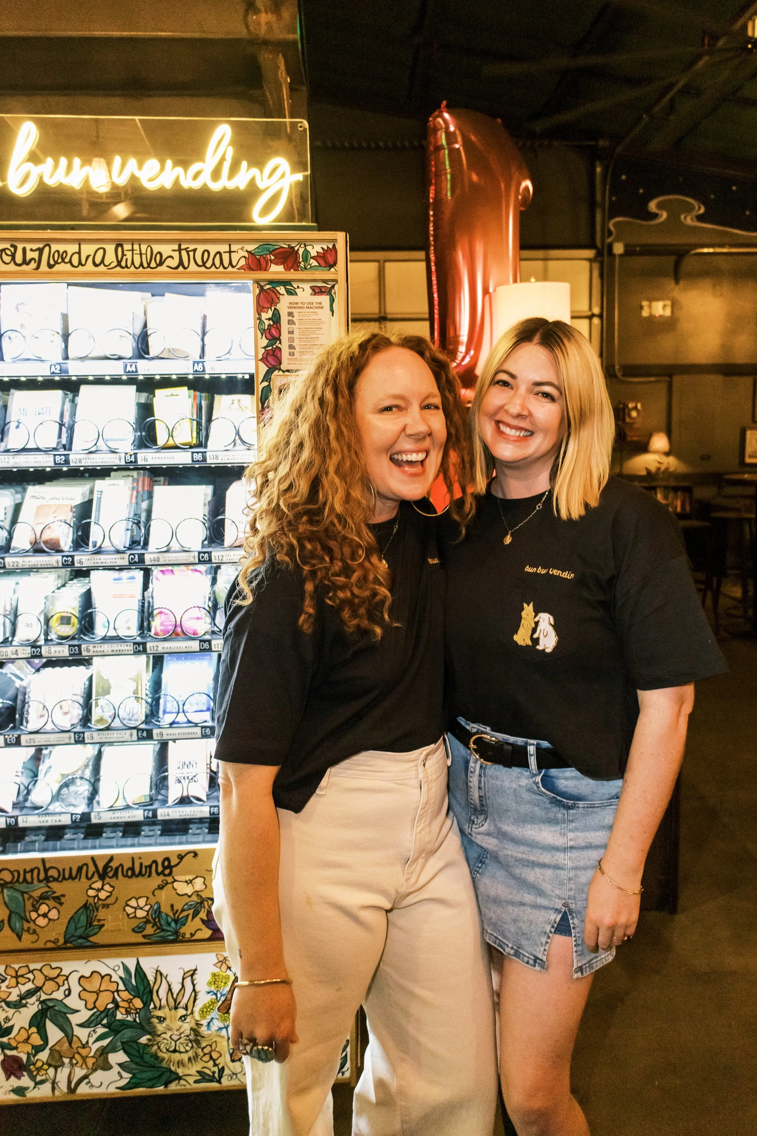 Two women smiling and standing close together in front of a vending machine at a bubble tea shop. One woman has long, curly red hair and is wearing beige pants, and the other has blonde hair and is wearing a black t-shirt with a dog graphic and a denim skirt. There is a neon sign that says 'bubvending' and decorative art with floral and animal designs behind them.