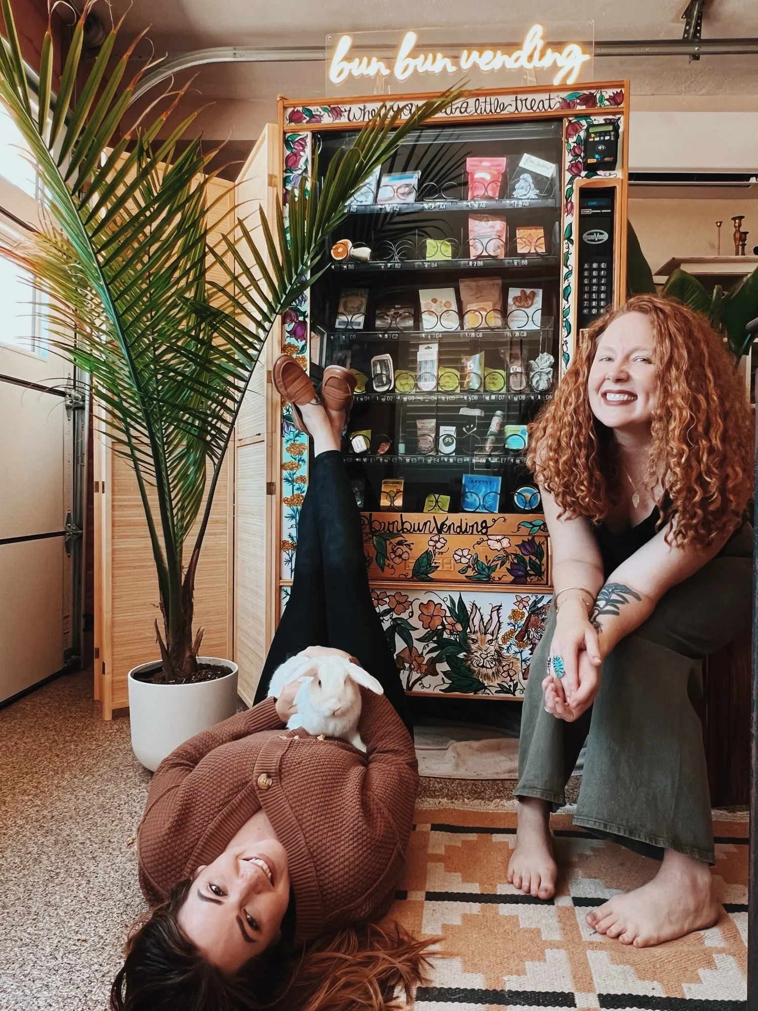 Two women  front of a vending machine with plants around. One woman is lying on the floor, smiling, with her head resting on her hand. The other woman is standing, smiling, holding the hand of the woman on the floor. The dog, a small white and brown breed, is lying on the ground near the woman on the floor. The vending machine displays various snacks and drinks. The setting appears to be indoors, possibly a cozy cafe or home area.
