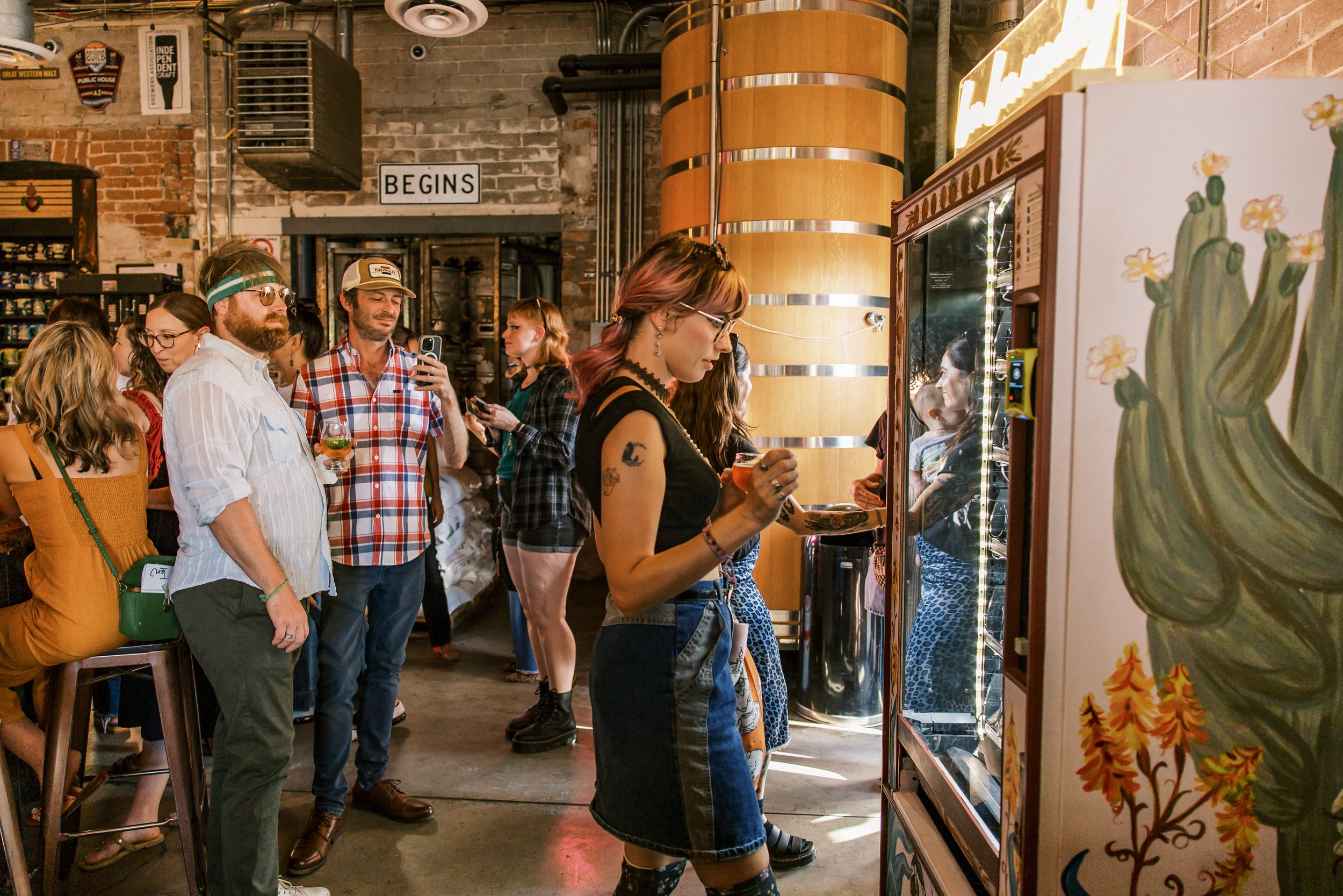 People gathered in a rustic indoor space, some observing a vending machine, with exposed brick walls, artwork, and a sign that says "BEGINS."