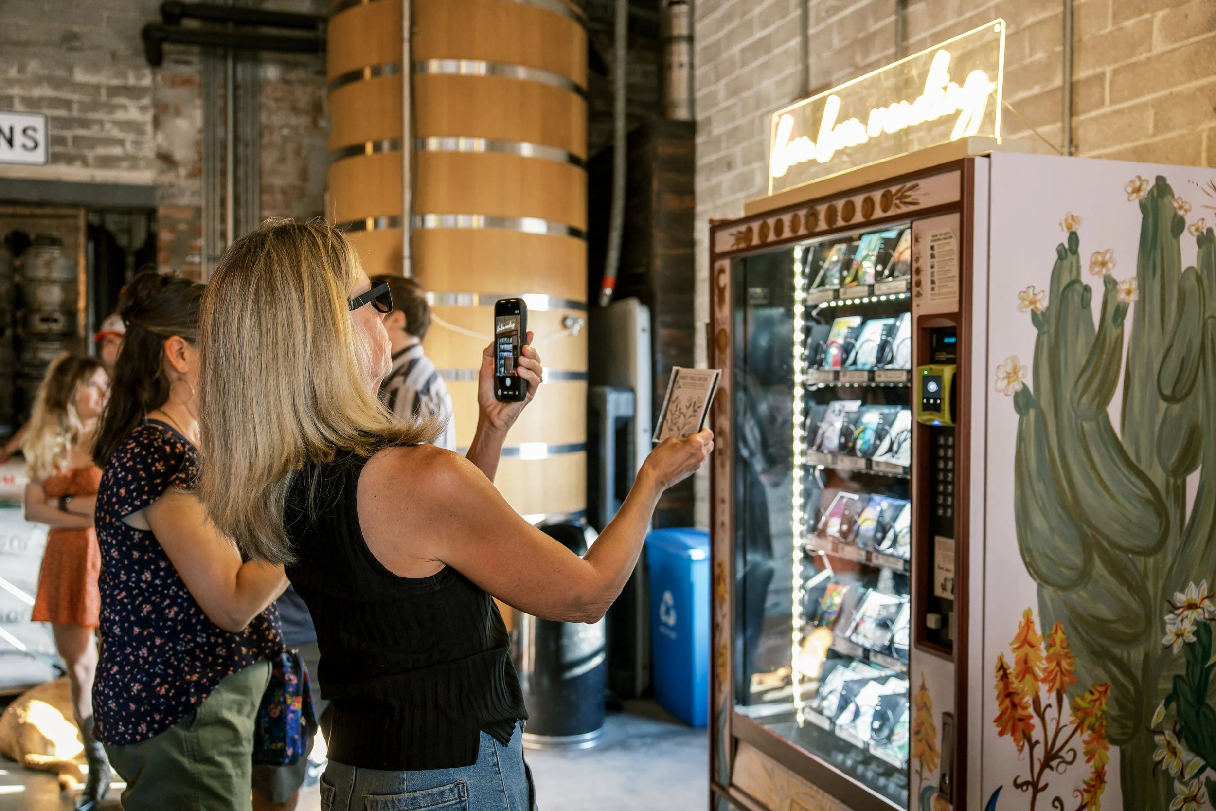 A group of people, mostly women, standing in line at a vending machine, with one woman taking a photo of the machine with her smartphone. The vending machine has floral and cactus artwork and is glowing, located inside a space with exposed brick walls and wooden barrels as decor.