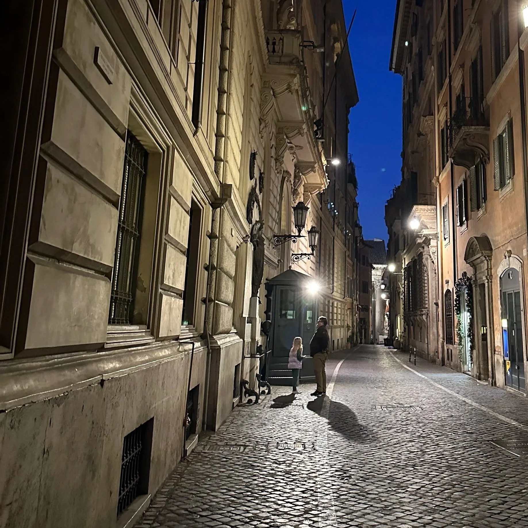 An empty cobblestone street in Rome in the predawn hours, softly lit by lamplight, with two people walking toward Trevi Fountain.