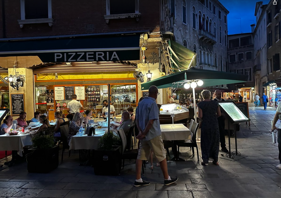 Outdoor diners at a Venice pizzeria on a brick-lined street at night, electric fans running against lingering heat.