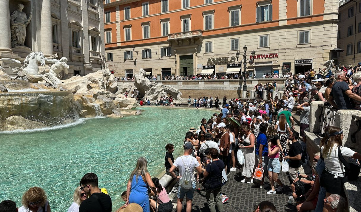 Crowds packed shoulder to shoulder around the Trevi Fountain in Rome on a sunny day, with tourists lining every available edge.